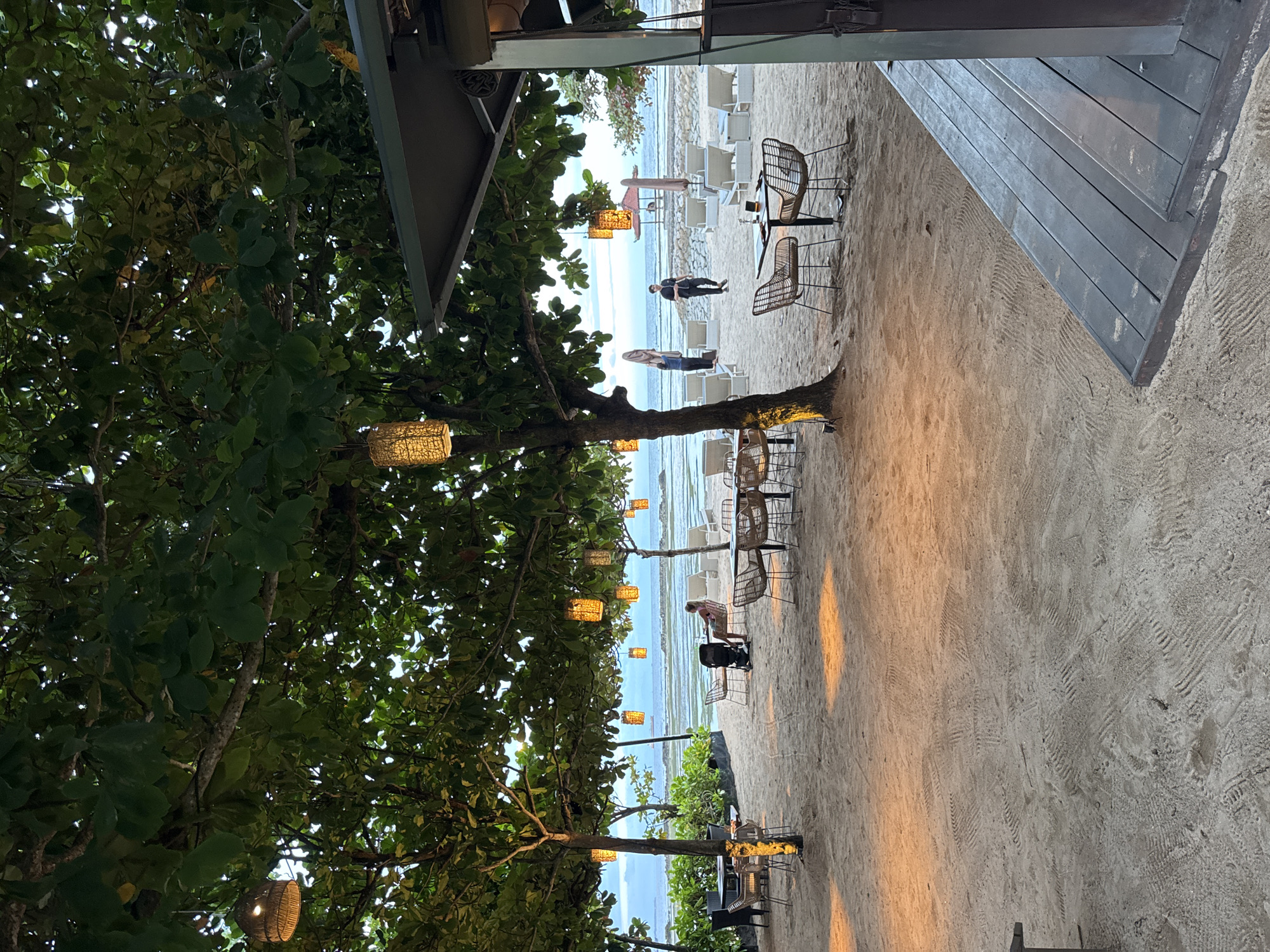 The image depicts an outdoor beach restaurant in Kuta Selatan, Indonesia. The view extends from beneath a shaded wooden deck area, across a sandy beach dotted with dining tables and chairs, towards the ocean. The foreground is dominated by a large tree adorned with decorative lanterns. Further back, some individuals stand or walk along the beach. Rows of lounge chairs are set up near the water's edge. The overall scene conveys a relaxed, coastal dining setting.