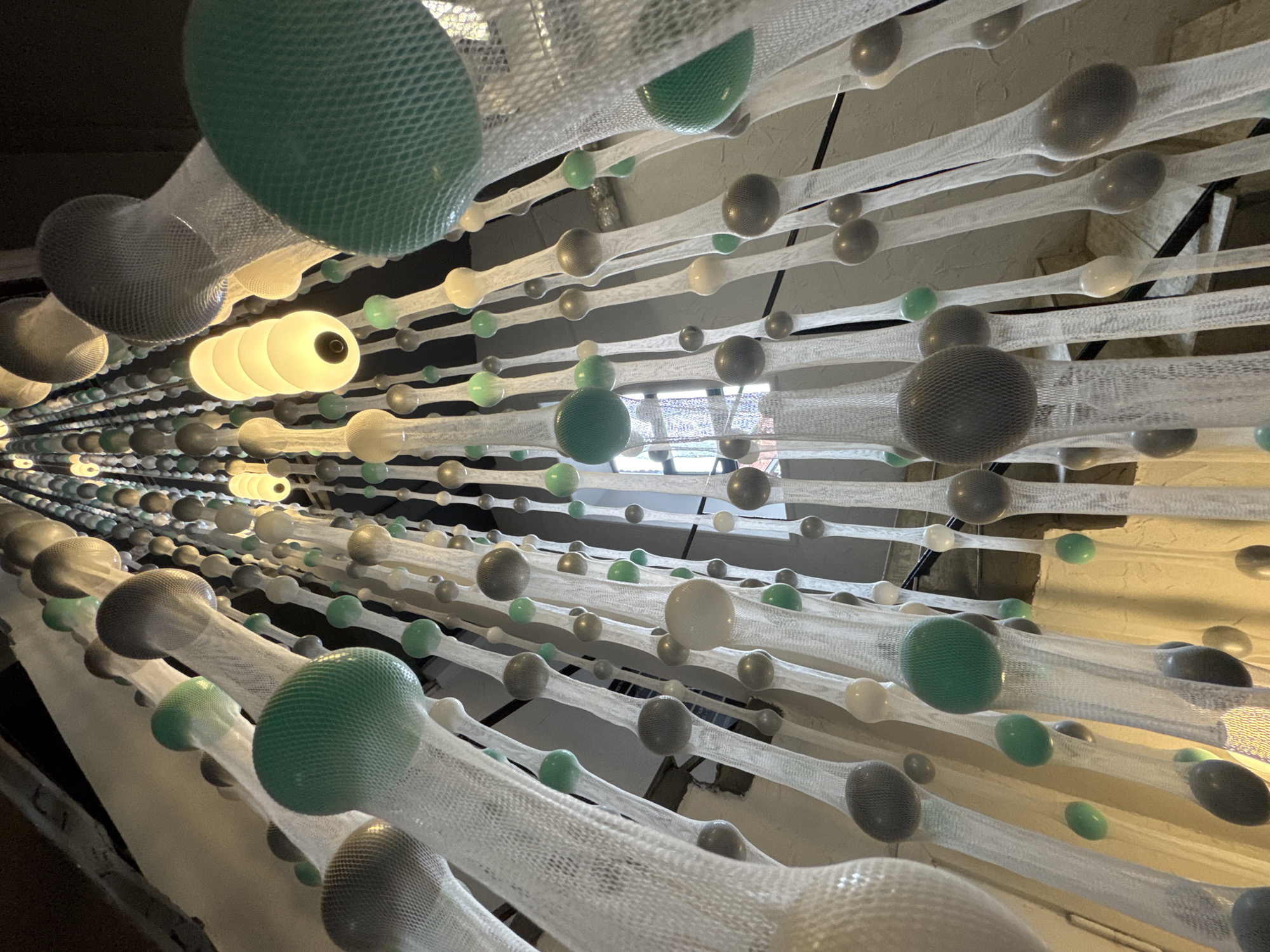 The image shows an abstract installation in an interior space, likely in Saint Petersburg, Russia. Rows of spheres, varying in size and color (green, gray, white), are suspended in white netting. The netting is taut, creating a horizontal alignment. Some spheres appear to be illuminated from within. Glimpses of the exterior are visible through a window in the background. The overall impression is of a modern art piece or architectural element.