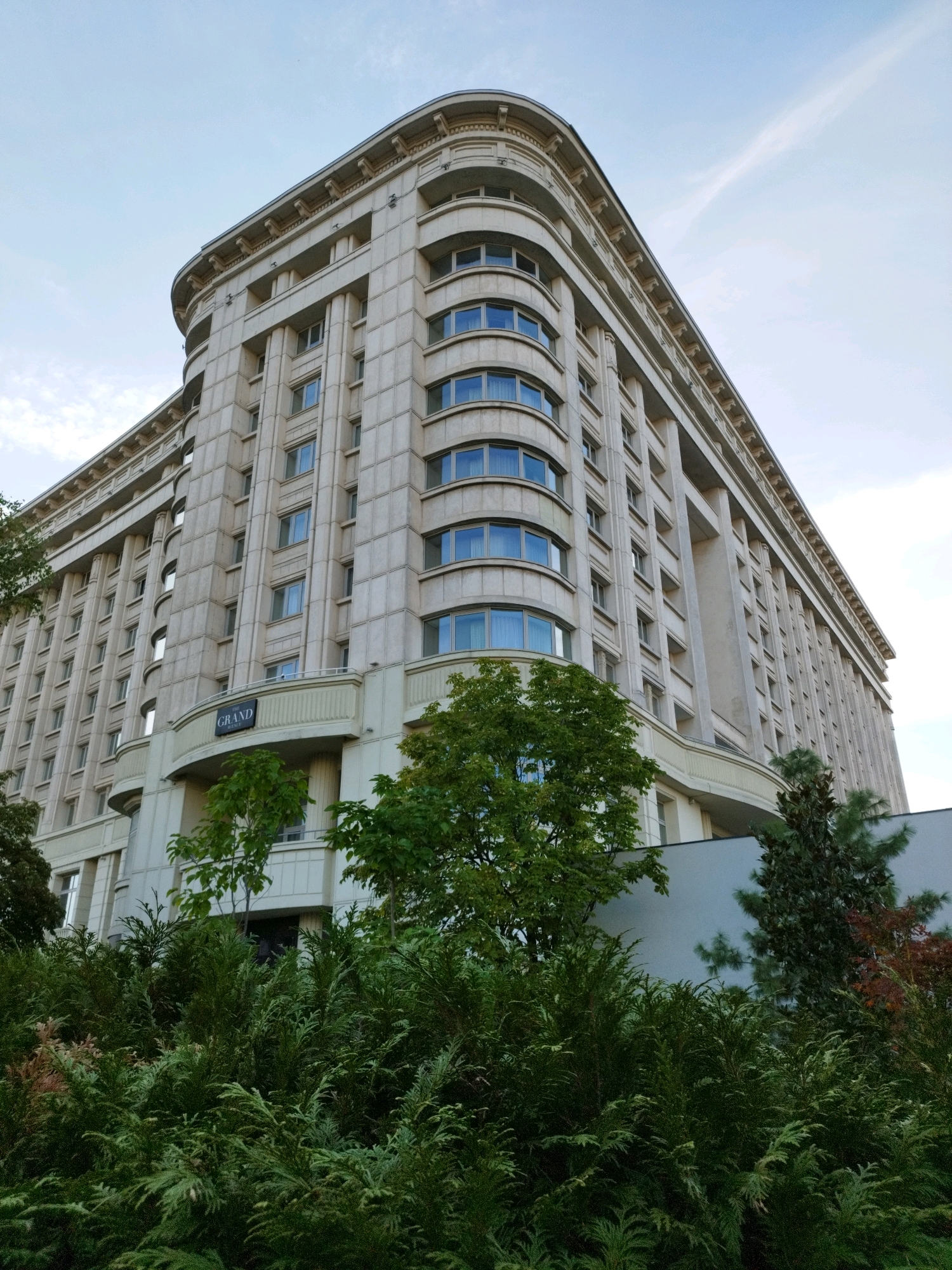 This is a low-angle, full shot of the Grand Hotel in Bucharest, Romania, taken from a street level perspective. The hotel, a large, beige, multi-story building with a curved facade, dominates the frame. Its architecture features repetitive vertical columns and numerous windows, many of which are arched. Above the entrance, a sign reads "GRAND HOTEL". In the foreground, a dense arrangement of lush green trees and bushes obscures the lower part of the building, with some autumnal foliage visible on the right side. The sky above is a pale blue with wispy white clouds. The scene is static and appears to have been captured during the daytime.