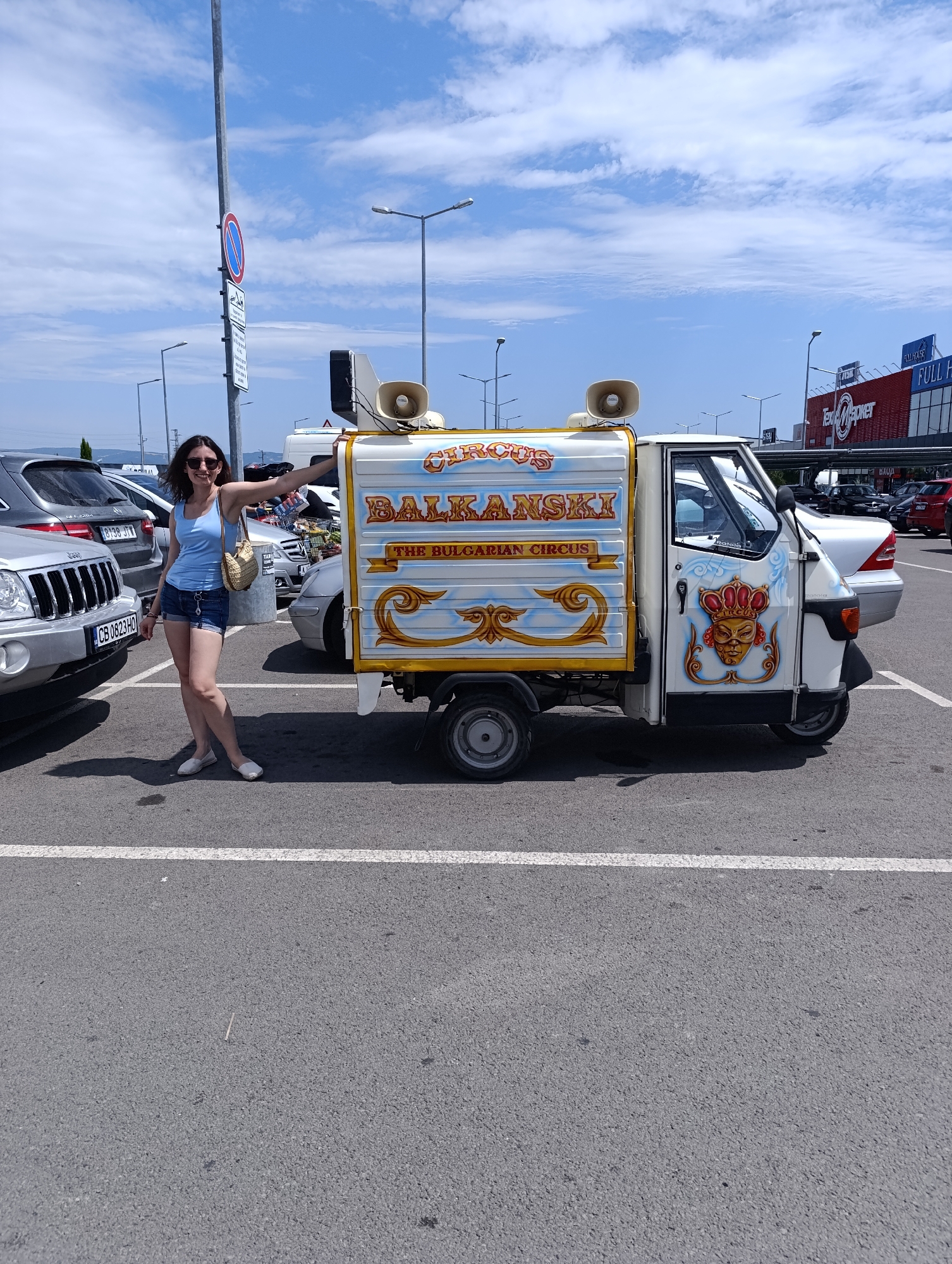 A woman stands next to a decorated three-wheeled vehicle in a parking lot. The vehicle, a white Piaggio Ape, is adorned with colorful graphics that include the words "CIRCUS BALKANSKI THE BULGARIAN CIRCUS" and an illustration of a crowned mask. The woman is wearing a blue tank top, denim shorts, and white slip-on shoes, and she is carrying a straw-colored purse over her shoulder. She is smiling and looking towards the camera, with her right arm resting on the vehicle. The parking lot appears to be in an urban area, with other cars and buildings visible in the background. A large building with "FULL HOUSE" and "TexMarket" signage can be seen in the distance. The sky is blue with scattered clouds. The scene appears to be set in Ravda, Bulgaria.