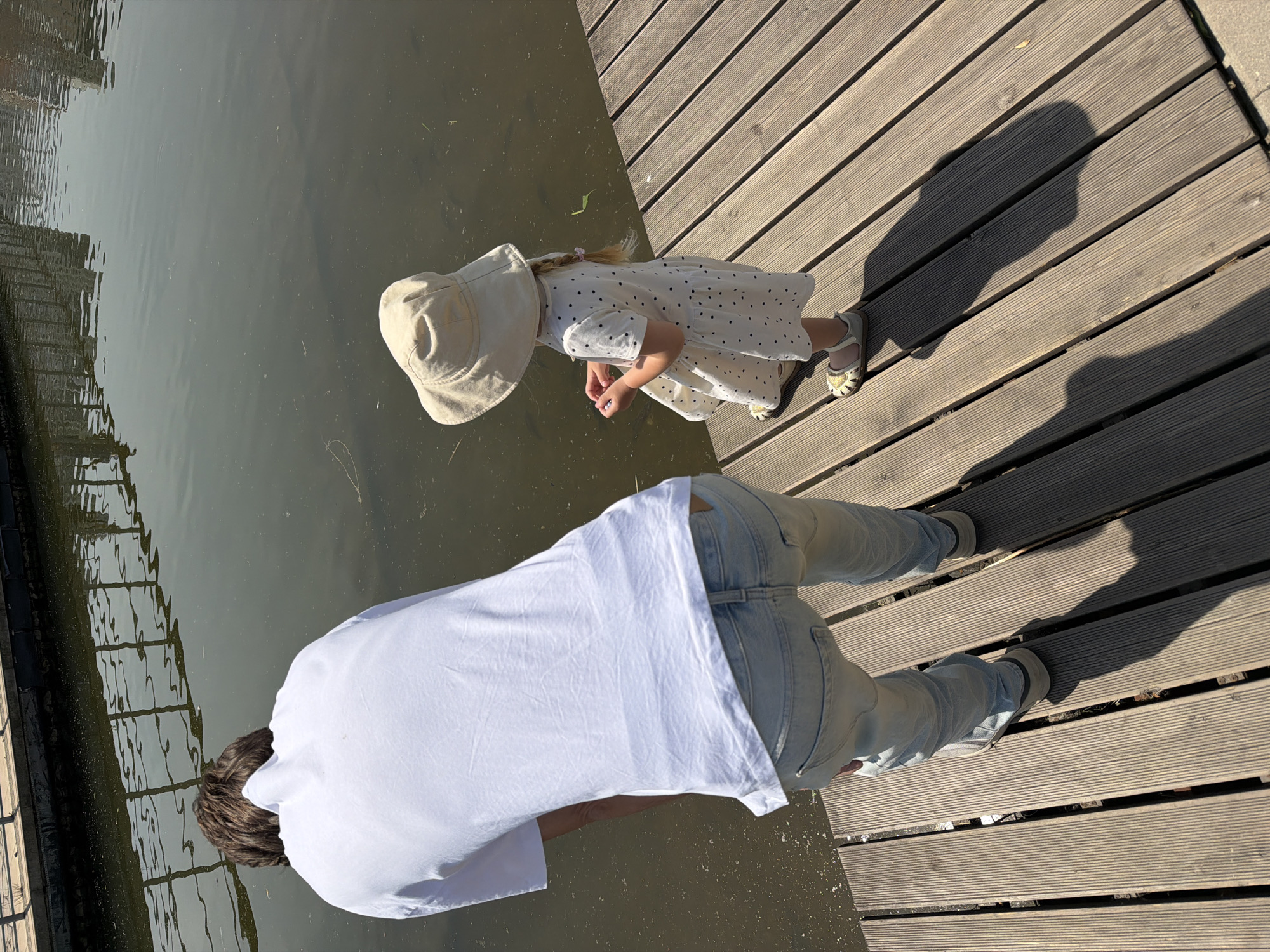 A high-angle view captures an adult and a child standing on a wooden boardwalk at the edge of a calm body of water in the city of Khimki, Russia.
The adult, positioned on the left, is seen from behind, leaning forward towards the water. They wear a white, short-sleeved t-shirt and light blue denim jeans. The t-shirt is wrinkled, and the waistband of light-colored underwear is visible above the jeans. Dark brown hair is visible on the back of the head and neck. Light-colored sneakers are visible on their feet.
To the adult's right, a young child stands upright, also viewed from behind, facing the water. The child wears a light beige, wide-brimmed sun hat, a white short-sleeved dress with dark polka dots, and light-colored sandals with yellow details. Blond hair, styled in a braid, extends from under the hat. The child holds a small, indistinct object in both hands at waist level.
The wooden boardwalk comprises multiple parallel, grooved, light brown planks. The water is dark and still, displaying reflections of overhead structures, possibly railings or trees, and scattered organic debris on its surface. Strong shadows of both individuals are cast onto the boardwalk, indicating direct sunlight from a high angle. Across the water, a distant bank features a low barrier or railing.