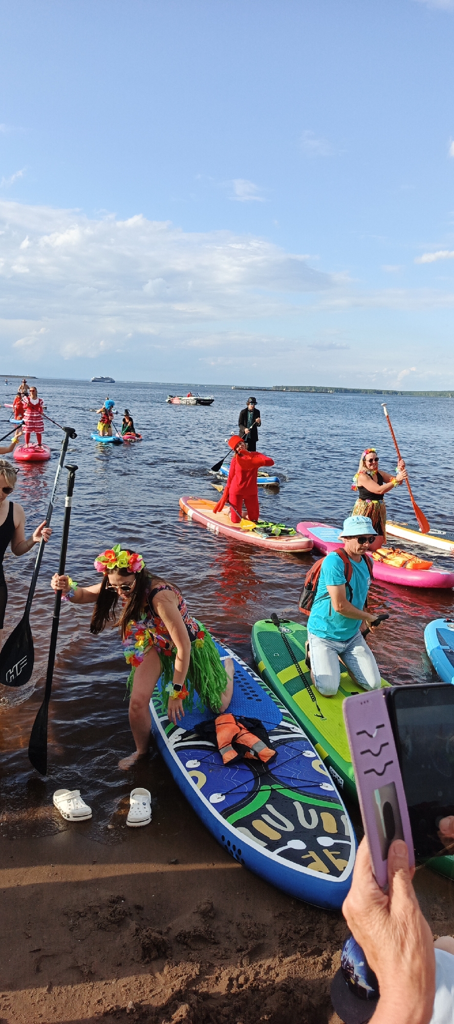 An outdoor daytime scene depicts a group of people on paddleboards in a body of water, likely a river or lake, near the shore. The sky is blue with scattered clouds. The water is dark with some reddish reflections. The shore is sandy and brown.
Several individuals are on paddleboards. One person in the foreground, wearing a floral headband and a hula skirt over a patterned top, is kneeling on a blue and yellow paddleboard. They are holding a paddle. Next to them on the sand are a pair of white Crocs. To their right, a person in a bright blue t-shirt and light-colored pants is kneeling on a green paddleboard, holding a paddle. Their face is obscured by a hat and sunglasses. In the middle ground, a person in a red full-body costume is standing on an orange paddleboard and holding a paddle. Further back, another person in a black suit and hat is standing on a paddleboard. Several other paddleboarders are visible in the distance, some in costumes including a red and white striped outfit and a yellow and black outfit. A small motorboat is visible in the distance. A large ferry or ship is on the horizon.
The scene appears to be a recreational event, possibly a paddleboard parade or gathering, with participants in various attire. The location is identified as the Kama River embankment in Chaikovsky, Russia, based on contextual information not explicitly present in the image itself.