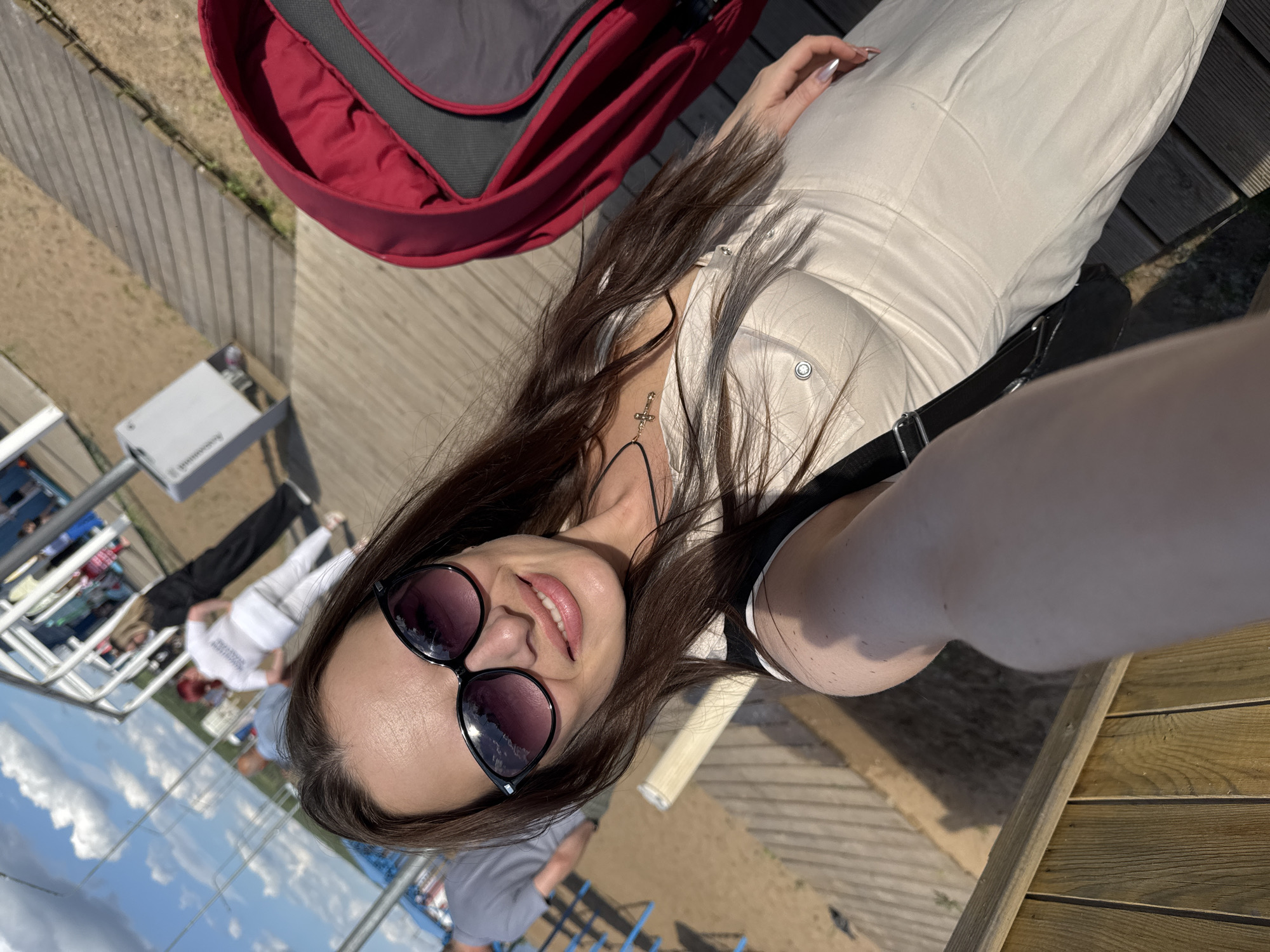 A woman with long brown hair and sunglasses is taking a selfie on a sunny day. She is wearing a light-colored dress and a black strap from a bag is visible. A red stroller is to her right. The background shows a wooden walkway, sand, a body of water, and people in the distance. The location is likely near the Kama River in Chaikovsky, Russia, possibly at a waterfront promenade or recreational area.