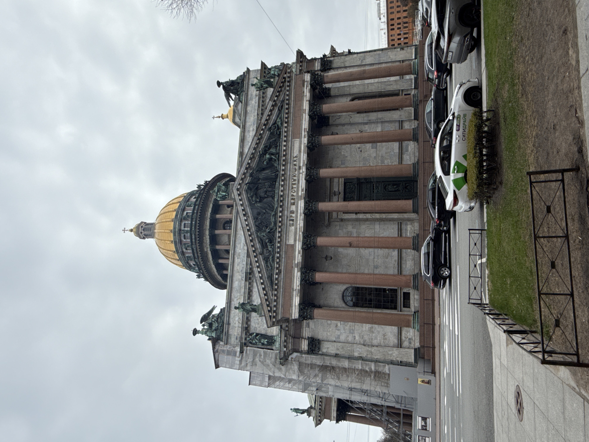 The image depicts St. Isaac's Cathedral in Saint Petersburg, Russia, captured on an overcast day. The grand neoclassical structure dominates the scene, featuring its distinctive golden dome, topped with a cross, and a massive portico supported by numerous reddish-brown granite columns. Intricate sculptures adorn the pediment above the columns, and a frieze below the pediment displays Cyrillic text, clearly showing "ВЕРУЮЩИХЪ" (the faithful), part of a larger historical inscription.
To the right of the cathedral, a street runs parallel to the building, with several cars parked along the curb. One white car, identifiable as a car-sharing vehicle, features the green-and-white "СИТИДРАЙВ" (CITIDRIVE) logo and text, along with the number "1". A grey paved sidewalk with a black metal fence separates the street from a strip of green grass. The sky is uniformly grey and cloudy, indicating an overcast daytime setting with no direct sunlight. No people are visible, and the scene appears calm and static. The photograph is presented rotated 90 degrees clockwise from its natural upright orientation.