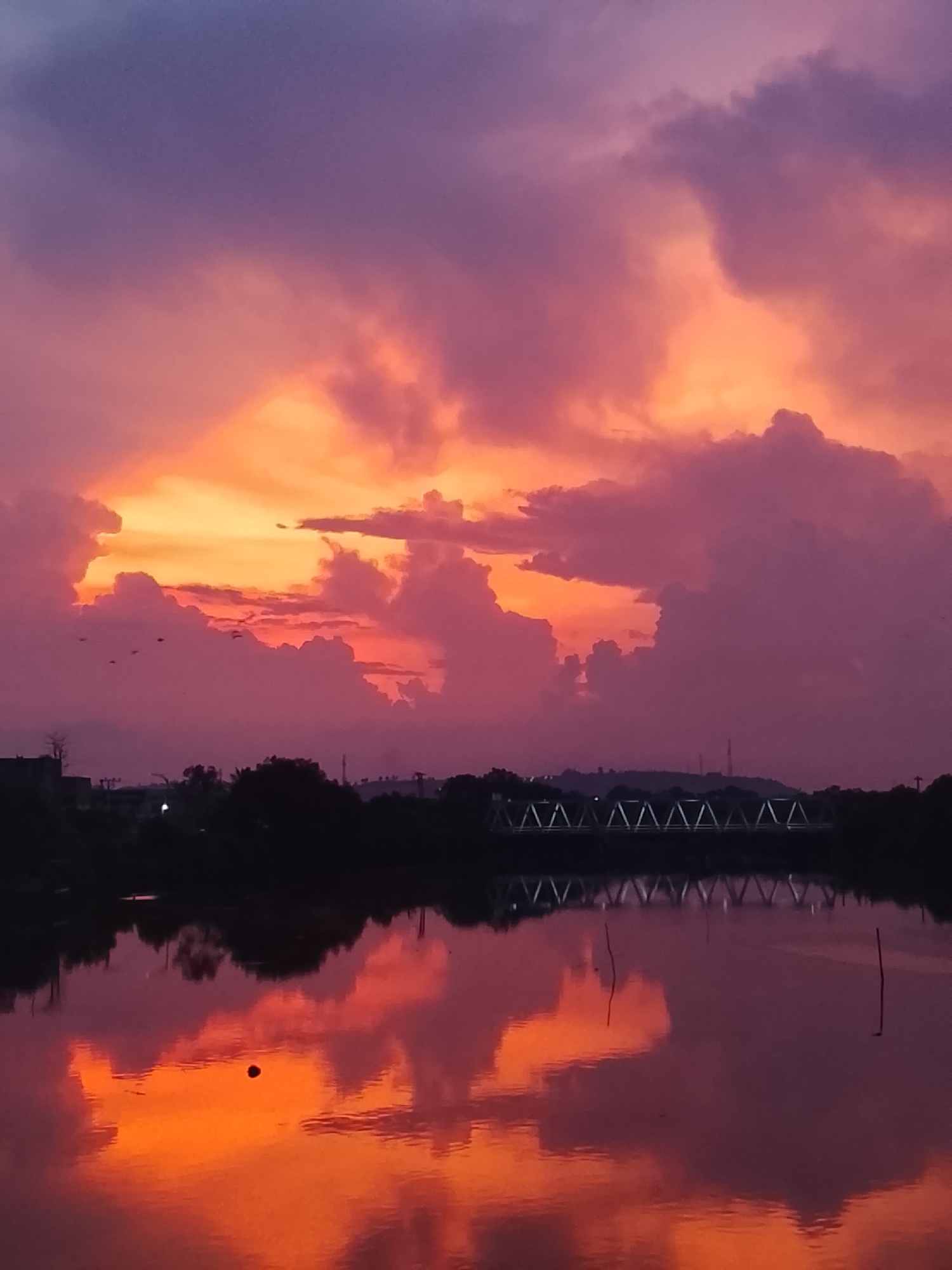 The image captures a breathtaking sunset or sunrise over a calm body of water in Lhokseumawe, Indonesia. The sky is a spectacle of dramatic clouds, intensely painted with vibrant hues of orange, yellow, pink, and purple. The colors are most brilliant near the horizon, gradually transitioning to deeper purples and blue-greys towards the top of the frame.
A dark, silhouetted landmass of trees, buildings, and possibly distant hills or mountains stretches across the horizon, separating the glowing sky from the reflective water. A prominent, light-colored truss bridge with a distinctive triangular pattern is visible against this dark silhouette in the middle distance.
The tranquil water surface acts as a near-perfect mirror, reflecting the spectacular sky and its colorful clouds with remarkable clarity and intensity. The reflections are slightly darker and more saturated than the actual sky. In the foreground water, a small, dark spherical object floats, and two slender poles are visible on the right side. A small flock of birds can be seen flying near the horizon on the left side of the frame.
The overall mood is serene and dramatic, showcasing the stunning natural beauty of a tropical twilight. No text is visible in the image.