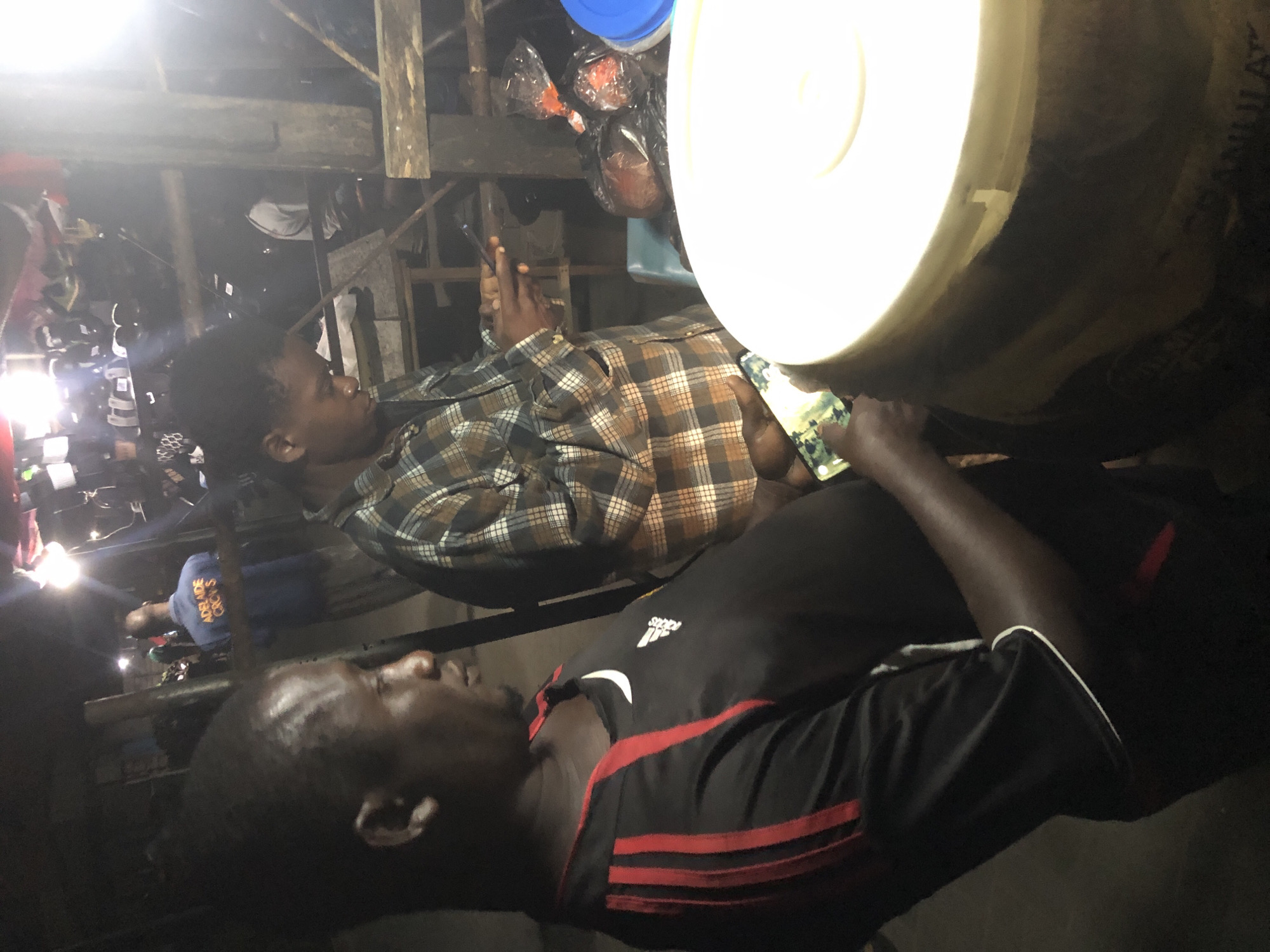 In Ado, Nigeria, at night, a man in a black Adidas shirt with red stripes is looking to his right. To his left, a person in a plaid shirt is holding a phone. In the background, another person wears a blue shirt with "ADELAIDE CREWS" written on it. The environment appears to be a market or roadside stall, with wooden structures and various items for sale, including food items in plastic bags and a large white container. The lighting is dim, with artificial lights creating a nocturnal atmosphere.