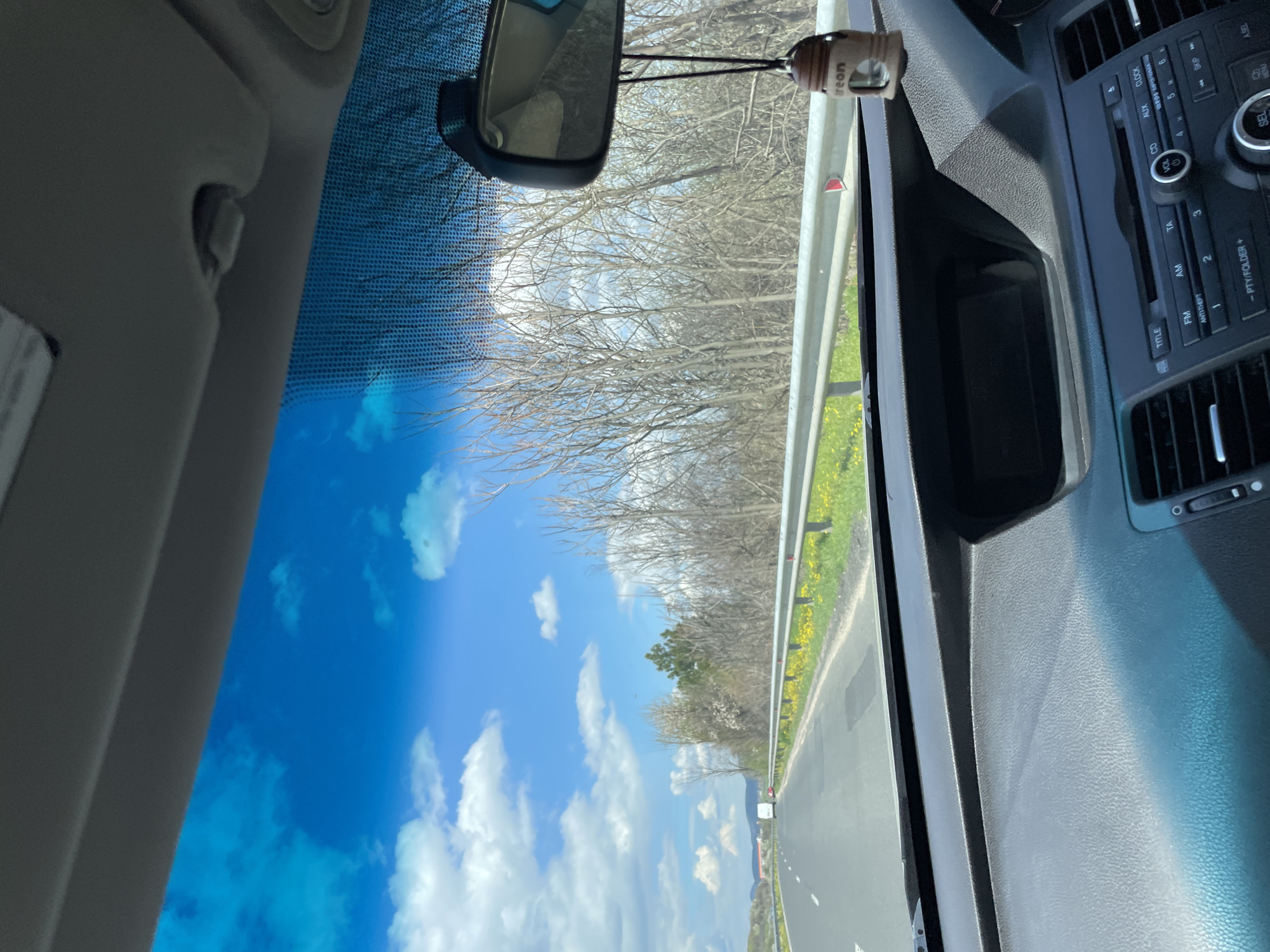 The image is taken from inside a vehicle, capturing a view through the windshield. Outside, a road stretches into the distance, bordered by a metal guardrail and a grassy area with yellow flowers. Beyond the road, leafless trees fill the horizon under a blue sky dotted with white clouds. Inside the vehicle, the rearview mirror is visible, along with the dashboard and radio console of the car. The radio console shows buttons and labels such as "FM," "AM," "CD," "AUX," "VOL," "CLOCK," and "ANTI-THEFT." An air freshener is hanging from the rearview mirror. The location, Stareishino, Bulgaria, is not confirmed by the image.