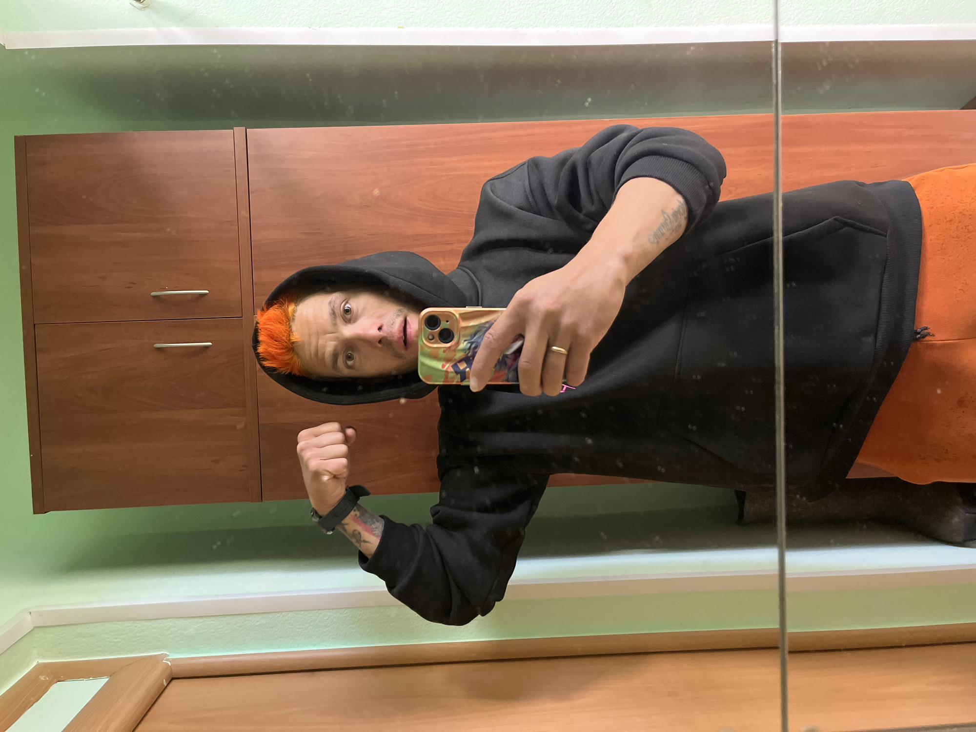 A man with dyed orange hair, wearing a black hooded sweatshirt and orange pants, is taking a selfie in front of a mirror. He is holding a phone in his right hand, flexing his left arm, and looking at the camera with a surprised expression. The background includes wooden cabinets, light green walls with white trim, and a wooden door. The reflection in the mirror shows the man's attire and some details of the room. The location is Chaikovsky, Russia.