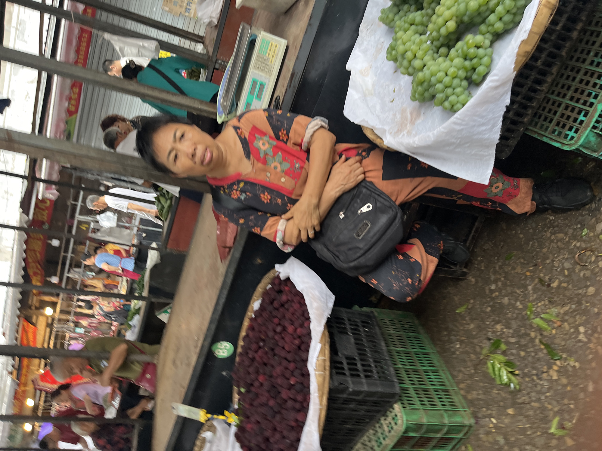The image depicts a bustling market scene in Jiangjin, China. In the immediate foreground, a woman is seated on the right, facing slightly left. She wears a patterned long-sleeved shirt and matching trousers in hues of orange, black, and red, and carries a black shoulder bag. Her arms are crossed. To her right, a display of green grapes, resting on white paper within a tray, is visible atop green plastic crates. Behind her, a silver and green digital weighing scale sits on a dark counter.
To the left of the seated woman, a large circular tray holds a substantial quantity of dark red, round fruit, possibly bayberries or lychees, also resting on white paper. This tray is stacked on two dark plastic crates, which are on a green plastic crate. A yellow price tag holder is attached to the counter above the fruit display, alongside a green sticker marked "B5."
The midground and background reveal a vibrant market environment with numerous individuals and stalls. On the left, an older man with white hair, wearing a green polo shirt and a red cross-body bag, is visible behind the fruit display. Other figures in the background are engaged in market activities, with various stalls displaying produce and hanging cuts of meat. On the right side of the background, a person in a bright teal long-sleeved top with a black bag walks past. The market structure consists of metal support poles and an overhead covering. The ground is damp and shows scattered green leaves and debris, indicative of active commerce.