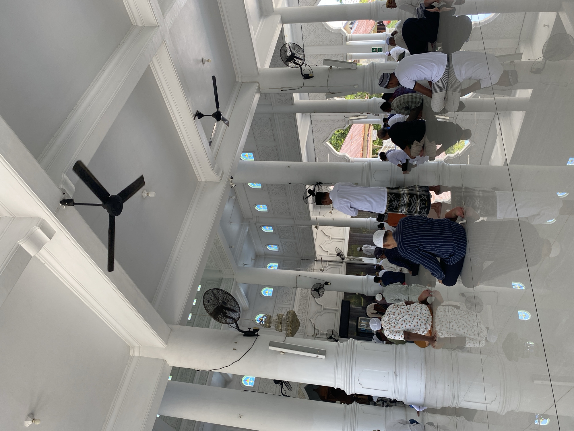 A group of men are praying in a well-lit mosque. The mosque has white columns, white beams, and ceiling fans. The floor is shiny and reflects the worshippers. The worshippers are kneeling on the floor. The location is Lhokseumawe, Indonesia.