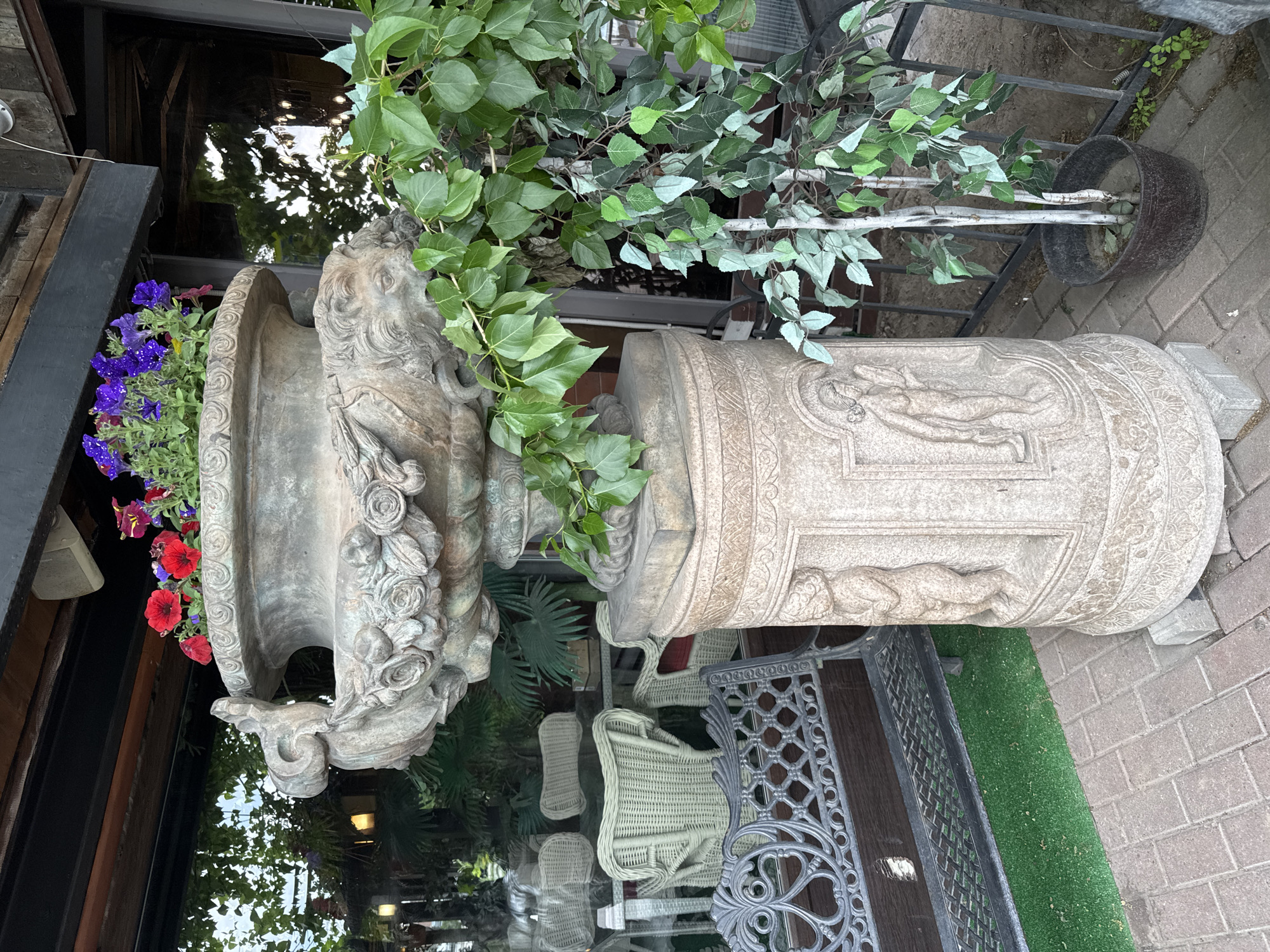 The image displays an ornate outdoor decorative arrangement, likely part of a building's entrance or patio. Two large, elaborate planters dominate the center of the frame under natural daylight.
On the left, a dark, weathered urn-shaped planter is richly adorned with intricate carvings, including what appear to be leonine heads, swirling floral patterns, and classical motifs. This planter holds a vibrant mix of flowering plants, predominantly purple petunias with some red blooms. To its right, a lighter-colored, cylindrical stone pedestal or planter stands, featuring detailed bas-relief carvings of draped figures and decorative borders.
Behind and between these heavy planters, a tall plant or artificial tree with light green, broad leaves grows upwards. Its base is contained within a simple dark pot resting on the ground.
The background shows a building facade with large, dark windows that reflect the sky, surrounding foliage, and hints of an interior or shaded outdoor area. Reflections in the windows reveal light-colored wicker chairs and an ornate dark metal table, suggesting an outdoor seating arrangement. The ground consists of reddish-brown brick pavement to the right, bordered by a strip of green artificial turf. Metal railings are visible in the distance.
No people are present, and no particular activity is depicted. The scene conveys a sense of classic outdoor ornamentation. No discernible text is visible in the image.