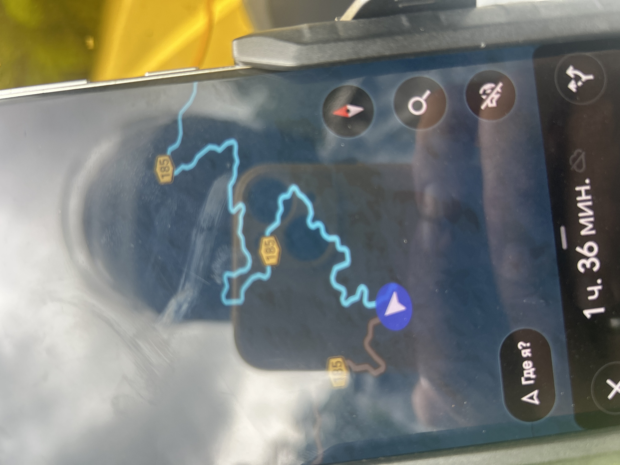 This image shows a smartphone screen displaying a navigation application. The application shows a map with a blue line indicating a route and several yellow markers with the number "185" on them, suggesting road numbers or points of interest. A blue arrow symbol indicates the current location or direction of travel. There are also several circular icons on the screen, including a compass, a magnifying glass for search, a symbol that may indicate a mute function, and a symbol representing a junction or turn. At the bottom of the screen, text displays "1 ч. 36 мин.", indicating a duration of 1 hour and 36 minutes. Below this, there is a button with Russian text "Где я?", which translates to "Where am I?". The overall environment appears to be a vehicle, as suggested by the reflections on the screen, the angle of the phone, and the yellow background visible through the phone's screen which could be a road or surrounding landscape. There is no discernible information within the image that specifically places it in Gopeng, Malaysia.