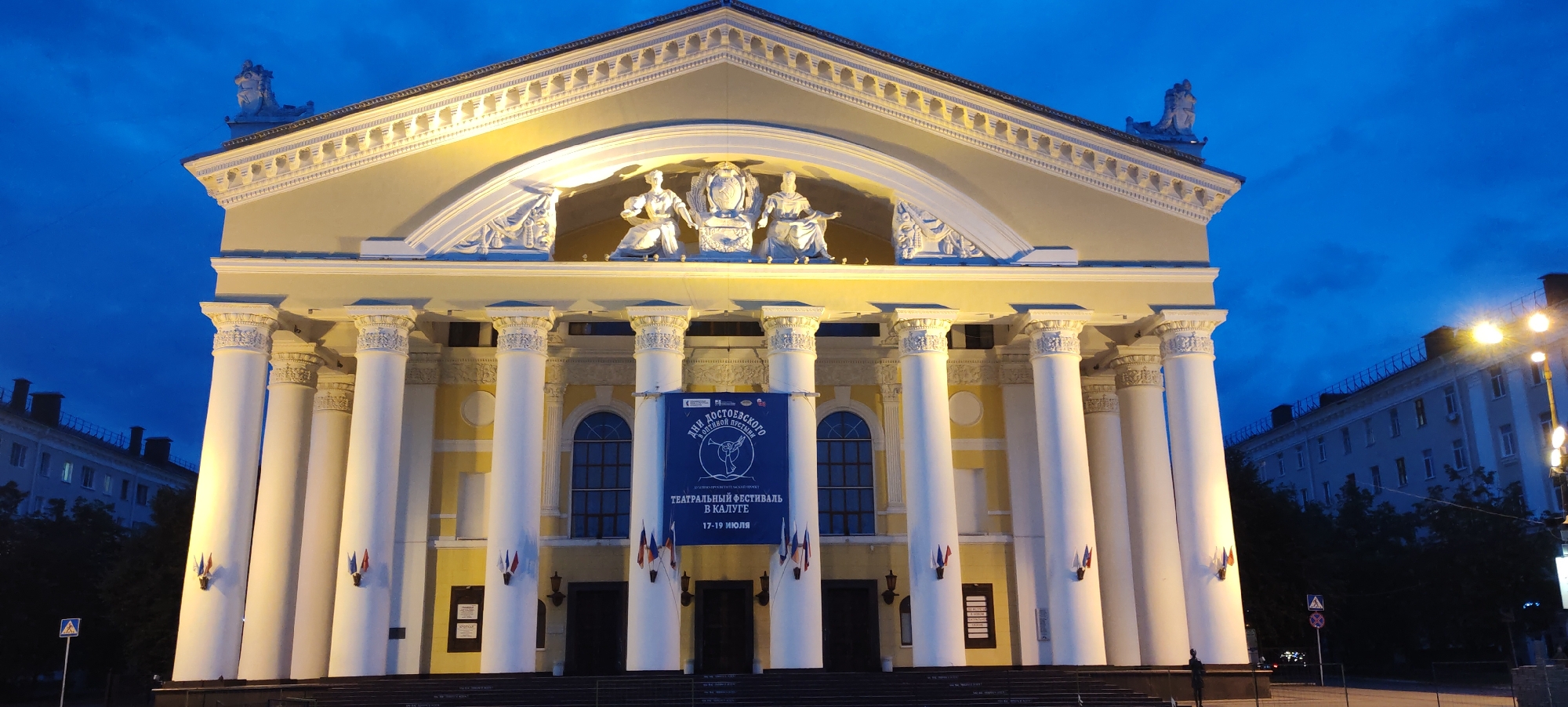 The image depicts the Kaluga Regional Drama Theatre, located in Kaluga, Russia. The theatre is a grand neoclassical building with a prominent facade featuring a pediment adorned with sculptures, a portico with large white columns, and decorative architectural elements. A large blue banner is hung across the facade, advertising the "Days of Dostoevsky" and the "Theatrical Festival in Kaluga" from July 17-19. Small flags are attached to some of the columns. The sky is a deep twilight blue, indicating the time is either early evening or late evening. Streetlights are visible on the right side of the image, illuminating the building and the surroundings. Trees line the street in front of the theatre. A pedestrian crossing sign and a traffic sign are visible on the left side of the image. A silhouette of a person can be seen standing on the steps in the lower right corner. The overall scene conveys a sense of cultural event and architectural grandeur.