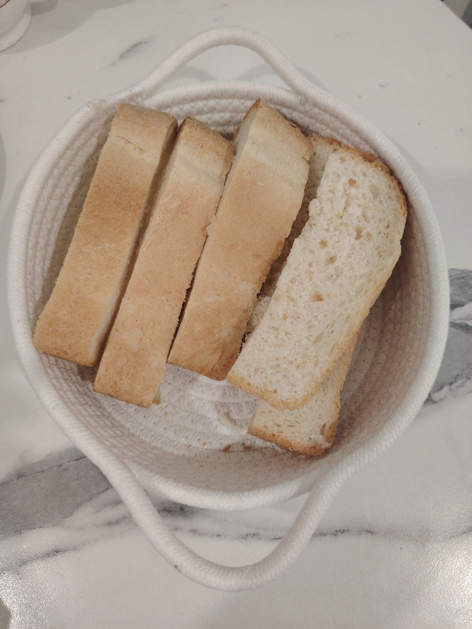 The image shows a white rope basket containing four slices of white bread. The bread slices are arranged vertically, with the two slices on the right showing more of the bread's interior. The basket has two rope handles and sits on a white surface with a marble pattern. The location in Sarapul, Russia is unknown from this image.