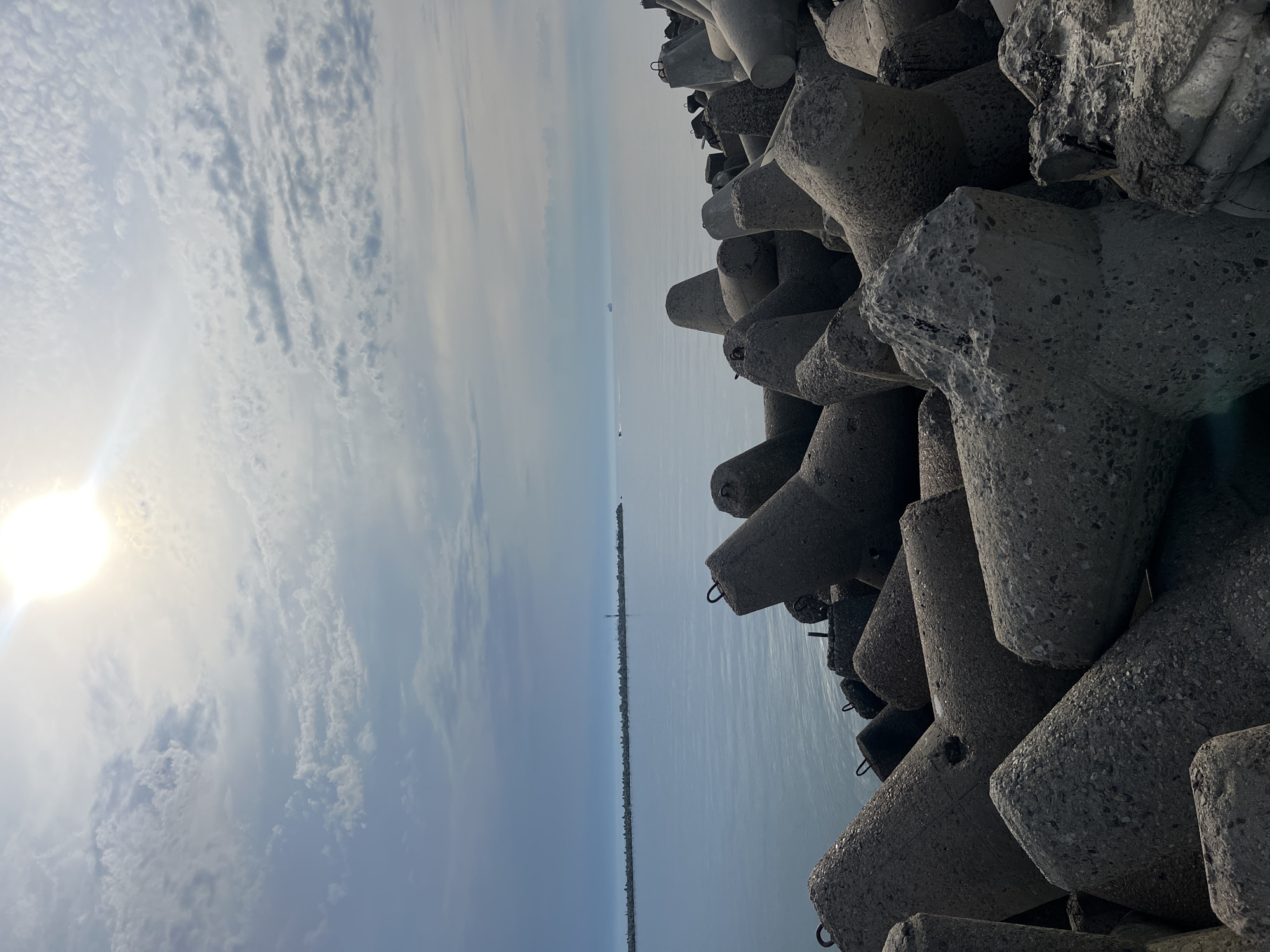 The image depicts a seascape with a breakwater constructed of concrete tetrapods in the foreground. The tetrapods are angular, grey, and appear weathered, stacked closely together. Beyond the breakwater, a calm, blue sea extends to the horizon, where a long, low line of breakwater structures forms a barrier, possibly a harbor entrance. In the distance, a small lighthouse or navigational marker is visible on this distant breakwater, and a few boats, including a larger ship, are present on the water. The sky above is partly cloudy, with the sun shining through, creating a bright area in the upper right quadrant of the image. The scene is likely located in Baltiysk, Russia, given the context of a Baltic Sea port.