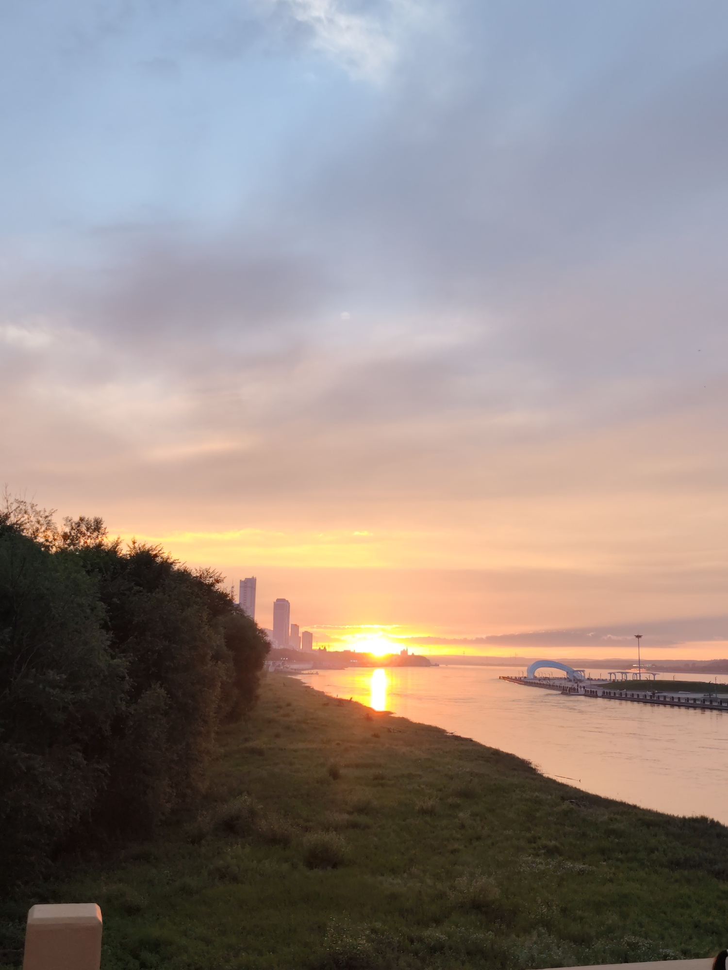 This is a landscape photograph taken at sunset, overlooking a wide river in 幸福乡, China. The sun is low on the horizon, casting a warm orange glow across the sky and reflecting brightly on the water. On the left side of the frame, a lush green hillside is covered in dense trees. Across the river, a city skyline with tall buildings is visible, silhouetted against the colorful sky. A modern structure, possibly a pier or a promenade with an archway, extends into the river on the right side of the image. The sky is a blend of soft blues and pinks, with scattered clouds. The overall scene is serene and picturesque, capturing the beauty of a coastal sunset.