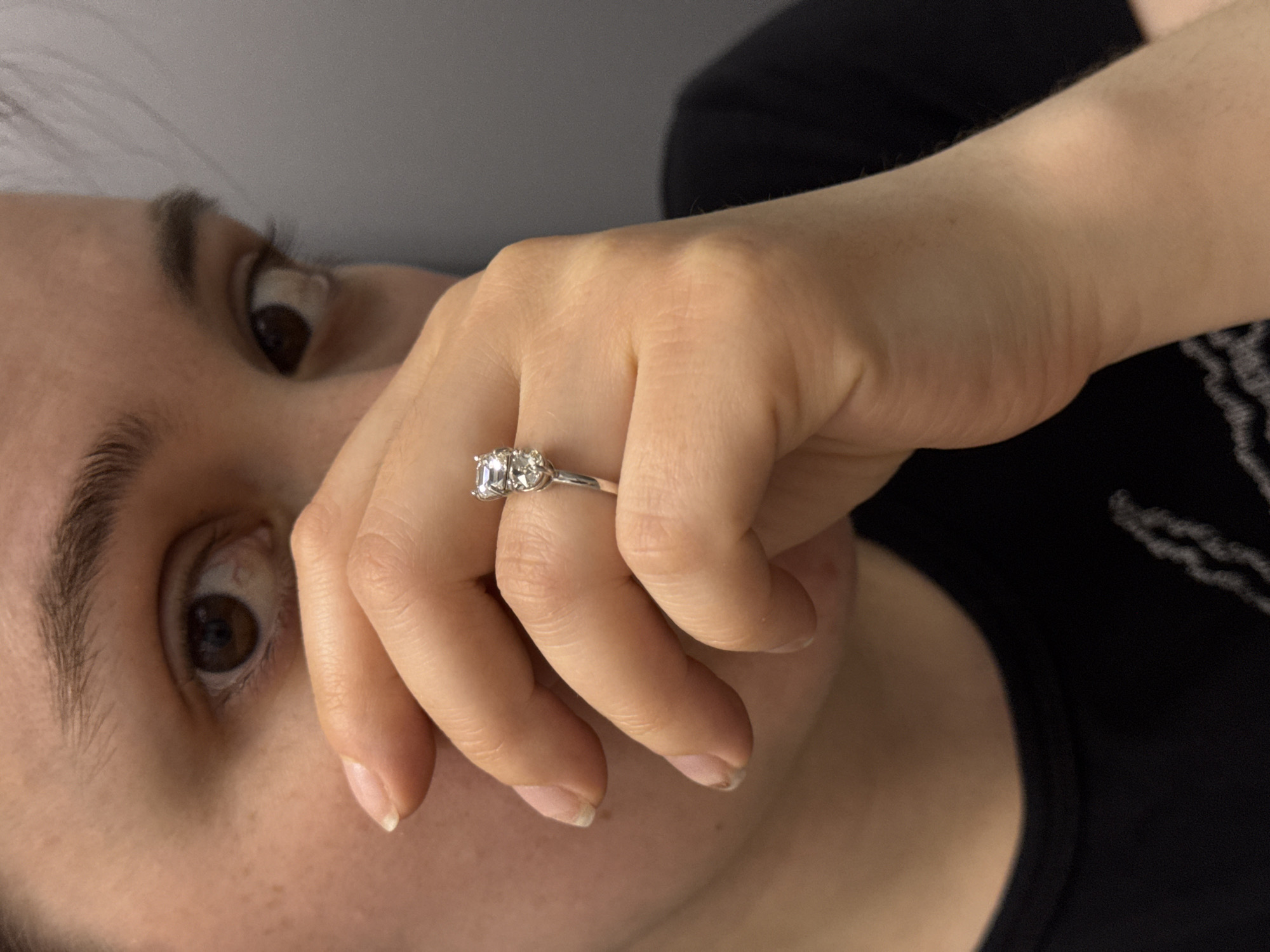A close-up image shows a person's face and hand with a three-stone ring on the ring finger. The person has fair skin, brown eyes, and visible freckles. Their hand is positioned in front of their face, partially obscuring their mouth. The ring features three square-cut diamonds set in a silver or white gold band. The background is a plain light gray wall and a portion of a black garment. There is no evidence to suggest that the image was taken in Izhevsk, Russia.