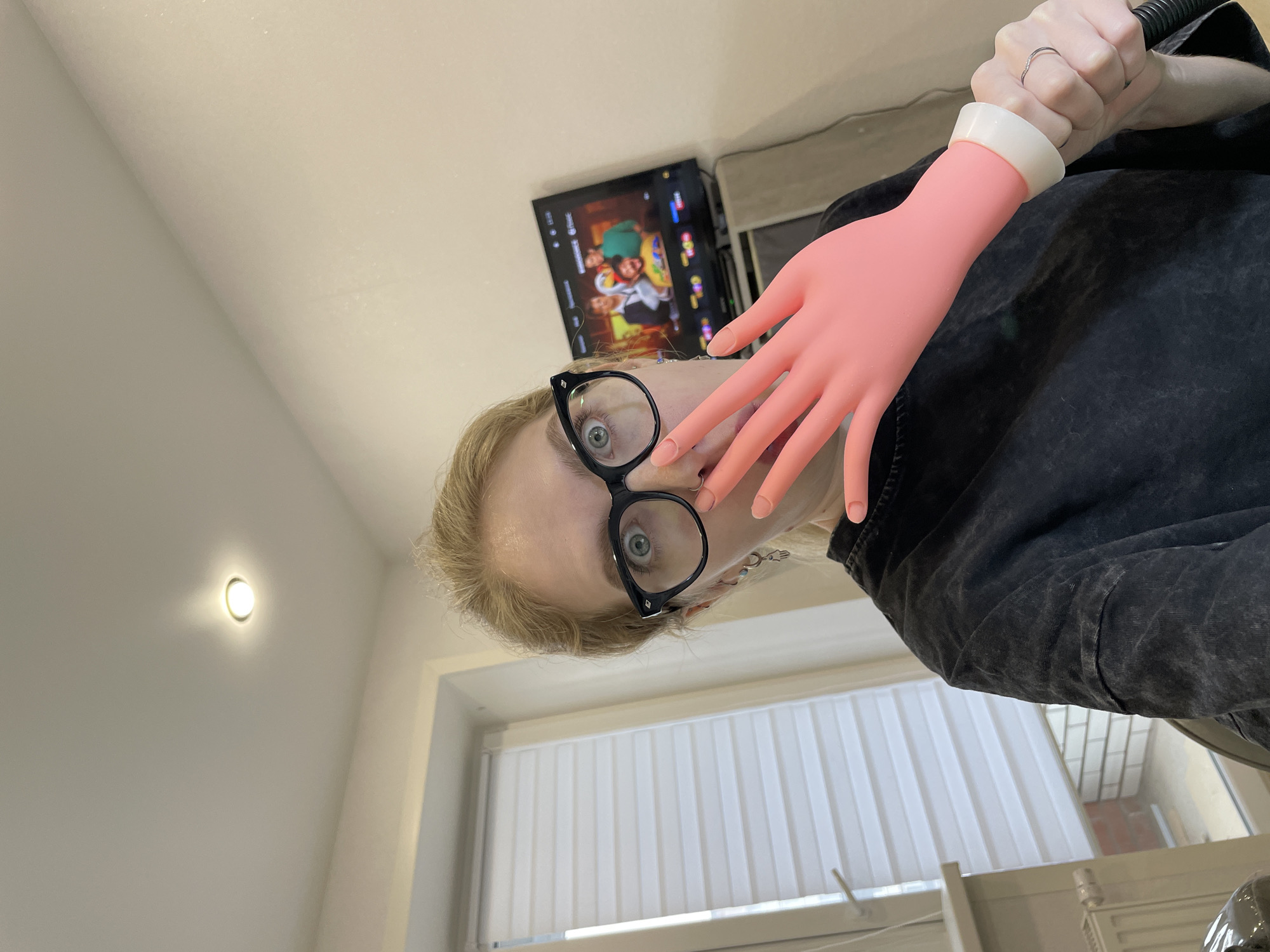 A tilted view of a person with glasses and a mannequin hand partially covering their face. The person has fair skin and blond hair. They are wearing black, large-framed glasses and a dark, textured garment. The mannequin hand is pink with a white wrist cuff and appears to be holding the nozzle of a vacuum cleaner.
Behind the person is a television displaying a cartoon or animated program. A window with white blinds is visible below the person. A round light fixture is on the white wall on the left.
The city of Izhevsk, Russia, cannot be identified in this image.