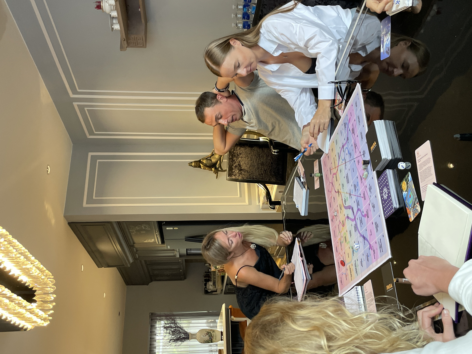 Four individuals are gathered around a glossy black table, engaged in what appears to be a board game. In the foreground, a woman with blonde curly hair is partially visible, her hands writing in a notebook. To her right, a woman in a white unbuttoned shirt, revealing a black top underneath, is holding a blue pen and also writing in a notebook. Her gaze is focused on the game. To her left, a man with short brown hair, wearing a grey t-shirt, leans back in his chair with his hands behind his head, looking towards the game. Facing the man, a woman with blonde hair, wearing a black dress, leans over the table, holding a pen and looking at a notebook. The table is covered with a colorful board game, dice, cards, and notebooks. The room features modern decor with grey walls, ornate trim, and a chandelier overhead. In the background, a kitchen area with dark cabinets and a refrigerator is visible, along with a window with vertical blinds and a vase with dried flowers. The setting is a modern, well-lit interior, possibly a dining room or living area.