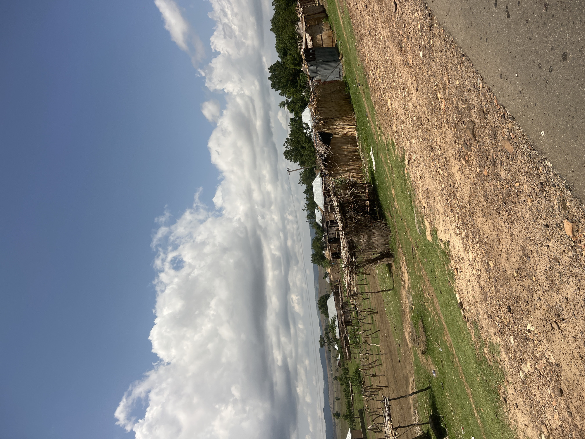 This image depicts a rural or semi-rural settlement in what is identified as Chum, Nigeria, under a partially cloudy blue sky. The scene is dominated by a collection of simple, rustic structures.
In the foreground, a paved dark gray road runs along the bottom right edge. Adjacent to the road is a wide, reddish-brown dirt and gravel shoulder or path, dotted with small stones and debris. A strip of vibrant green grass separates this unpaved area from the main village structures.
The midground features numerous dwellings or shelters, primarily constructed from natural materials such as dried grass, reeds, or thatch for walls and roofs. Several structures also incorporate corrugated metal sheets, particularly for roofing and some wall sections. These buildings are arranged in a linear fashion, suggesting a village layout. In an open area to the left of the main cluster of huts, there is a field containing multiple wooden stakes or poles driven into the ground, arranged in rows, indicative of agricultural activity or property demarcation.
Behind the structures, various trees with green foliage provide a natural backdrop, becoming denser on the right side of the image. Utility poles and power lines are visible horizontally across the mid-upper section, partially obscured by the trees. In the distant background, rolling hills or low mountains are visible beneath the horizon, appearing hazy. The sky above features a mix of bright white and darker gray cumulus clouds against a clear blue expanse. No people are visible within the frame.