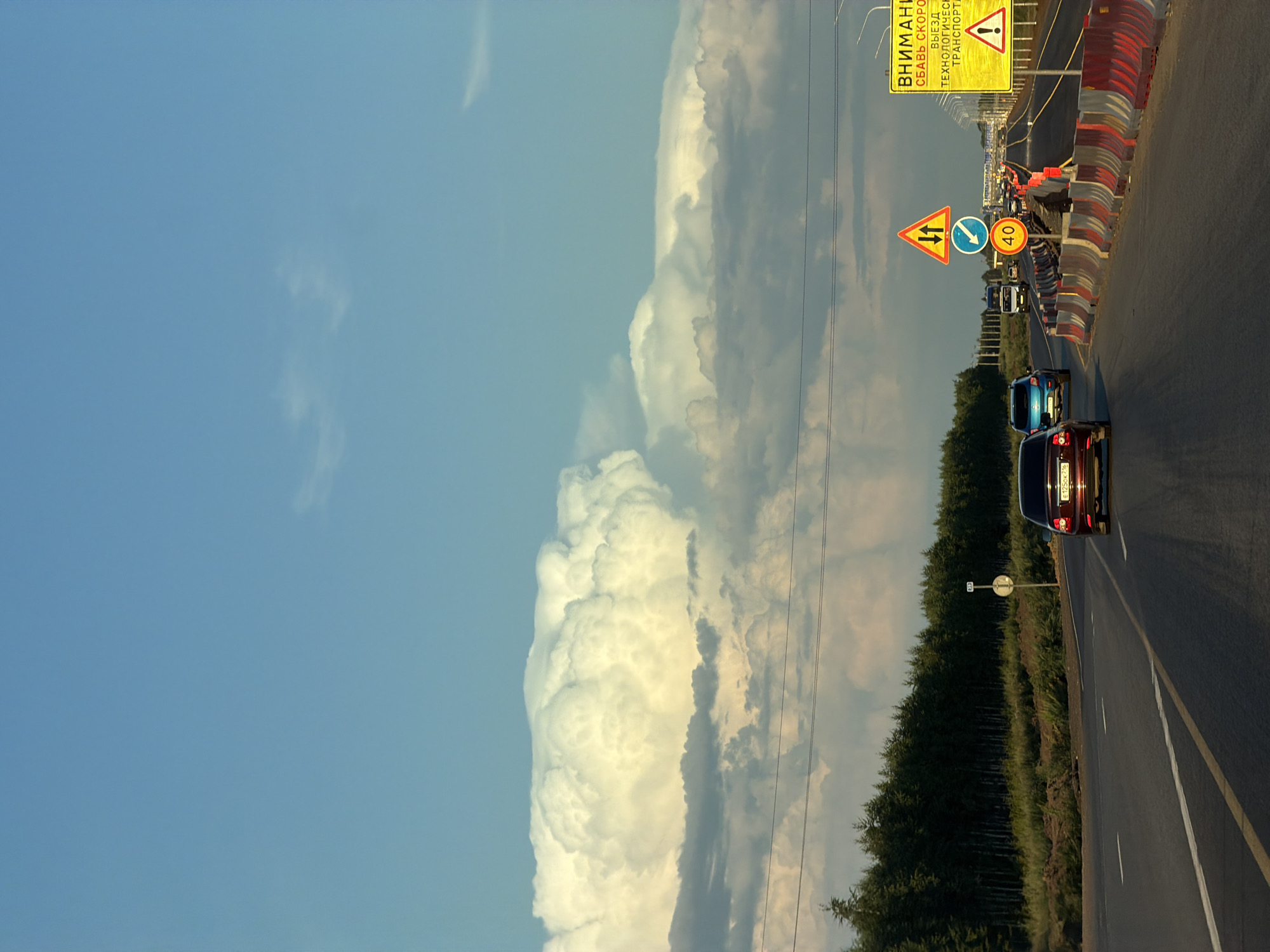 The image depicts a road with multiple lanes, bordered by a forest on the left and construction barriers on the right. Several cars are visible on the road, including a red sedan, a blue SUV, and a truck. Overhead, a massive, dramatic cloud formation dominates the sky, casting a warm light on the scene. Traffic signs are present, including a yellow warning sign with Russian text and a circular sign indicating a speed limit of 40. The overall impression is one of a journey through a rural or semi-rural landscape during late afternoon or early evening, with a prominent natural phenomenon in the sky. The road appears to be a highway or a major route, possibly undergoing some form of construction or maintenance due to the presence of barriers and traffic control signs.