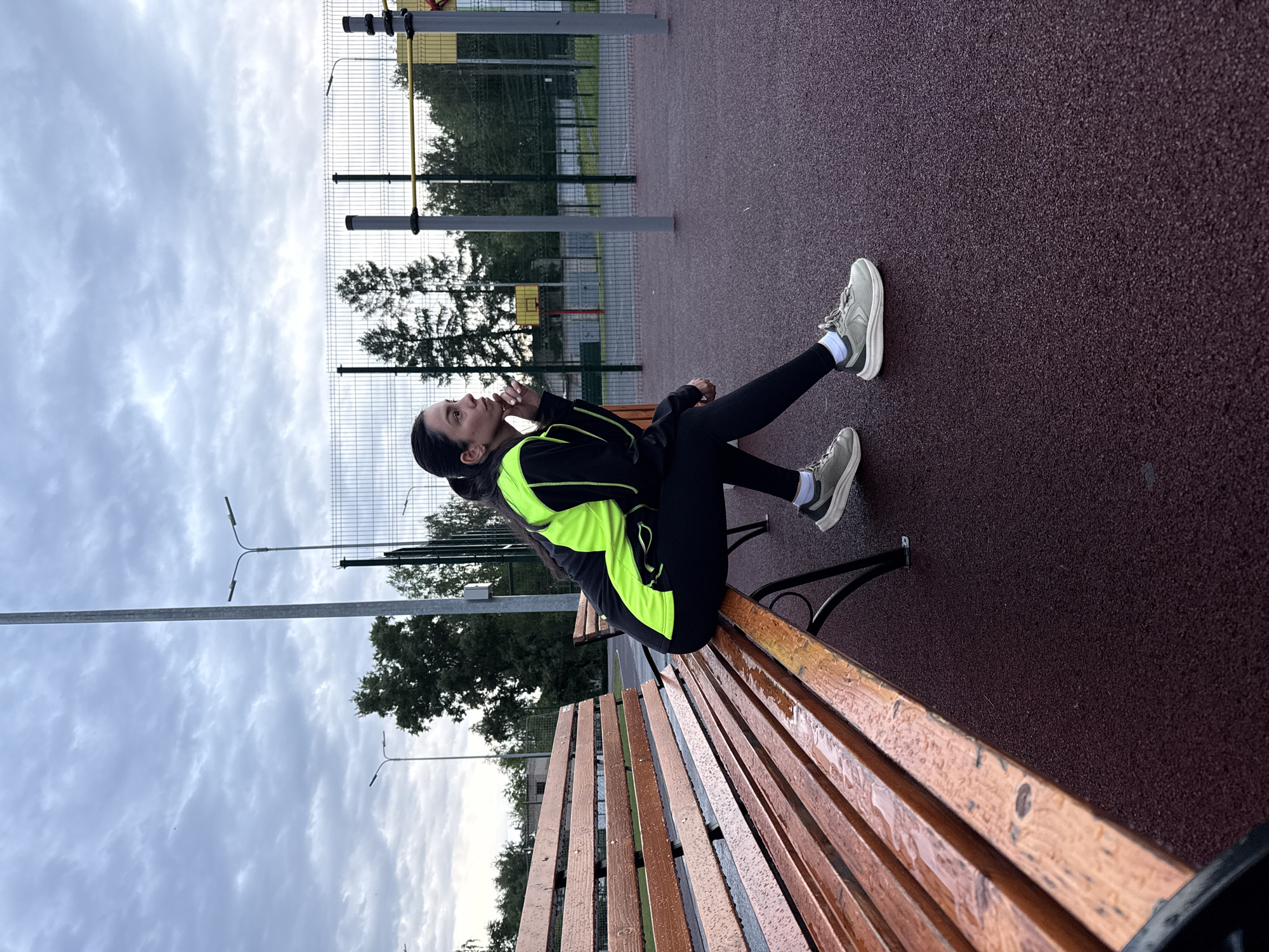 A woman is sitting on a wet wooden bench in an outdoor recreational area. She is wearing black leggings, grey sneakers, and a black jacket with neon green accents. She is looking upward with her hand near her face. The recreational area has a maroon rubberized surface, with workout equipment in the background, surrounded by a metal fence and green trees. Streetlights are visible in the background and the sky is overcast. The location is Chaikovsky, Russia.