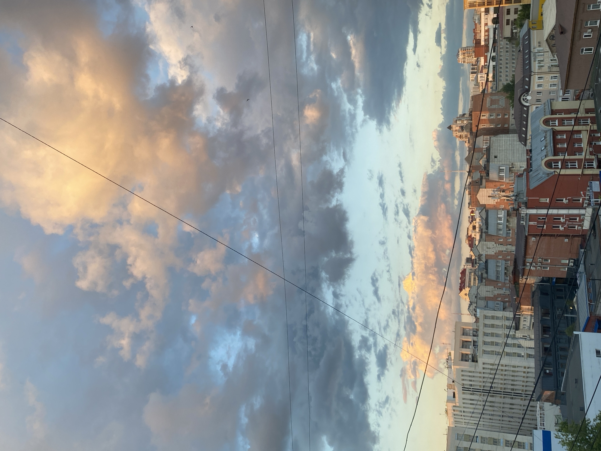 A cityscape is shown at sunset in Perm, Russia. The sky is filled with dramatic clouds, tinged with orange and pink from the setting sun, contrasting with areas of pale blue and dark gray. Several power lines crisscross the upper portion of the image. Below the sky, a dense collection of urban buildings is visible. The architecture varies, with some modern, tall structures featuring many windows, and others appearing older, with brick facades and traditional rooflines. Some buildings have visible air conditioning units. The overall impression is of a city viewed from a slightly elevated perspective during the golden hour.