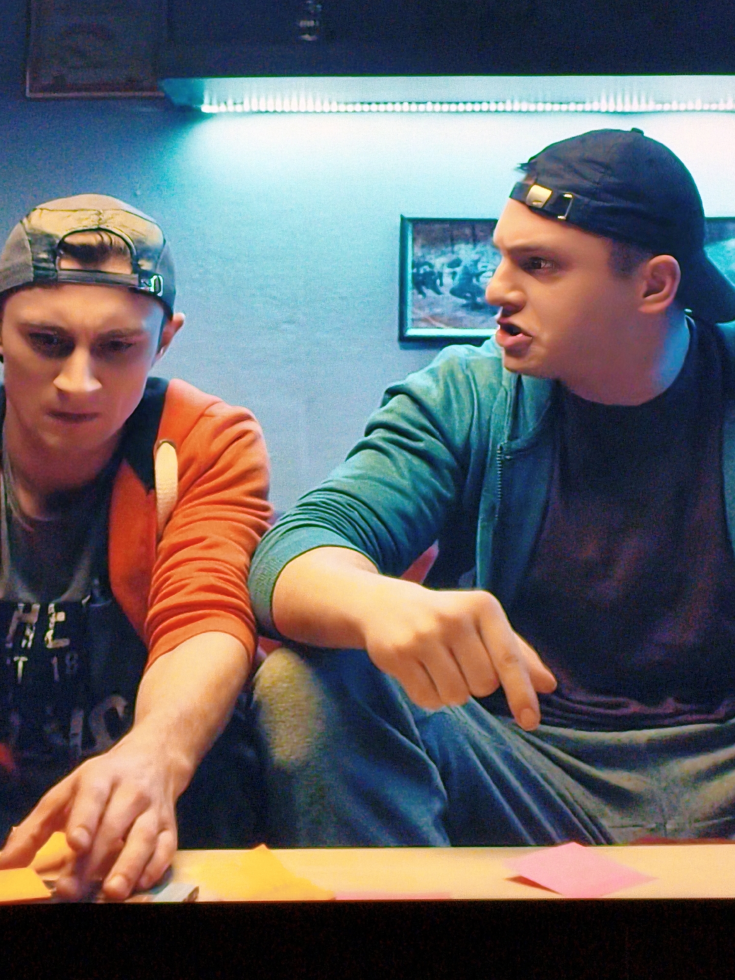 Two young men are sitting at a table, appearing to be engaged in a game or activity involving colored paper. The man on the left, wearing an orange jacket and a cap, is looking down at the table with a focused expression, his hands reaching out towards some yellow paper. The man on the right, wearing a blue jacket and a black baseball cap, is looking towards the left, speaking with an animated expression and pointing his index finger. The background suggests an indoor setting with a blue wall illuminated by a strip of white LED lights. A framed picture hangs on the wall behind the man on the right. The table is covered with colored paper, including yellow and pink. The scene does not provide any specific visual cues to indicate a location in Vladivostok, Russia.