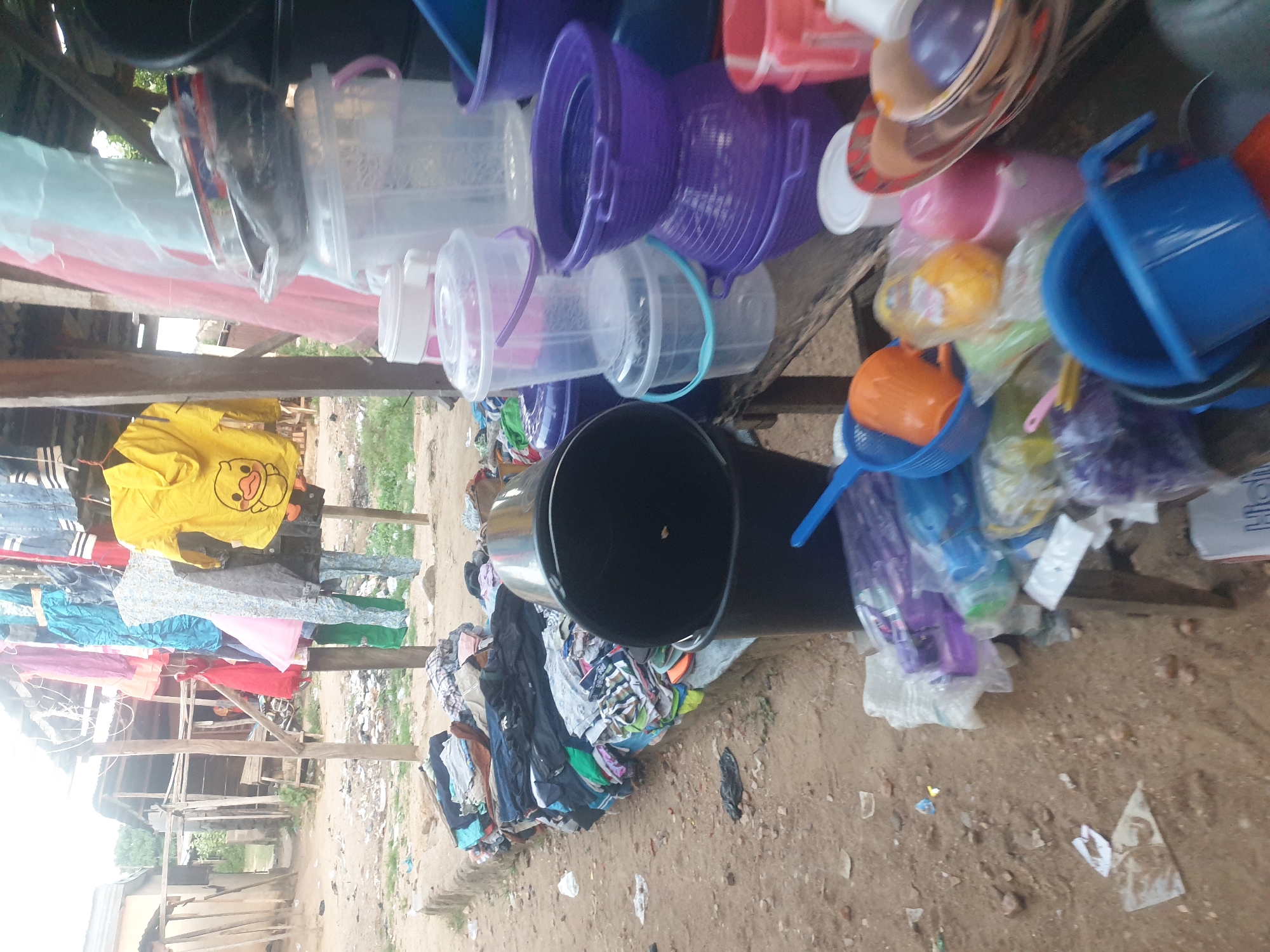 The image shows a vendor stall in an outdoor market in an urban environment, likely in Unknown. Plastic containers of various colors and sizes are displayed prominently along the top of the stall. Clothes hang from a makeshift clothesline strung across the stall frame. Below the containers, piles of folded and possibly used clothing lay on the ground, in what appears to be dirt and gravel. The area seems dusty.