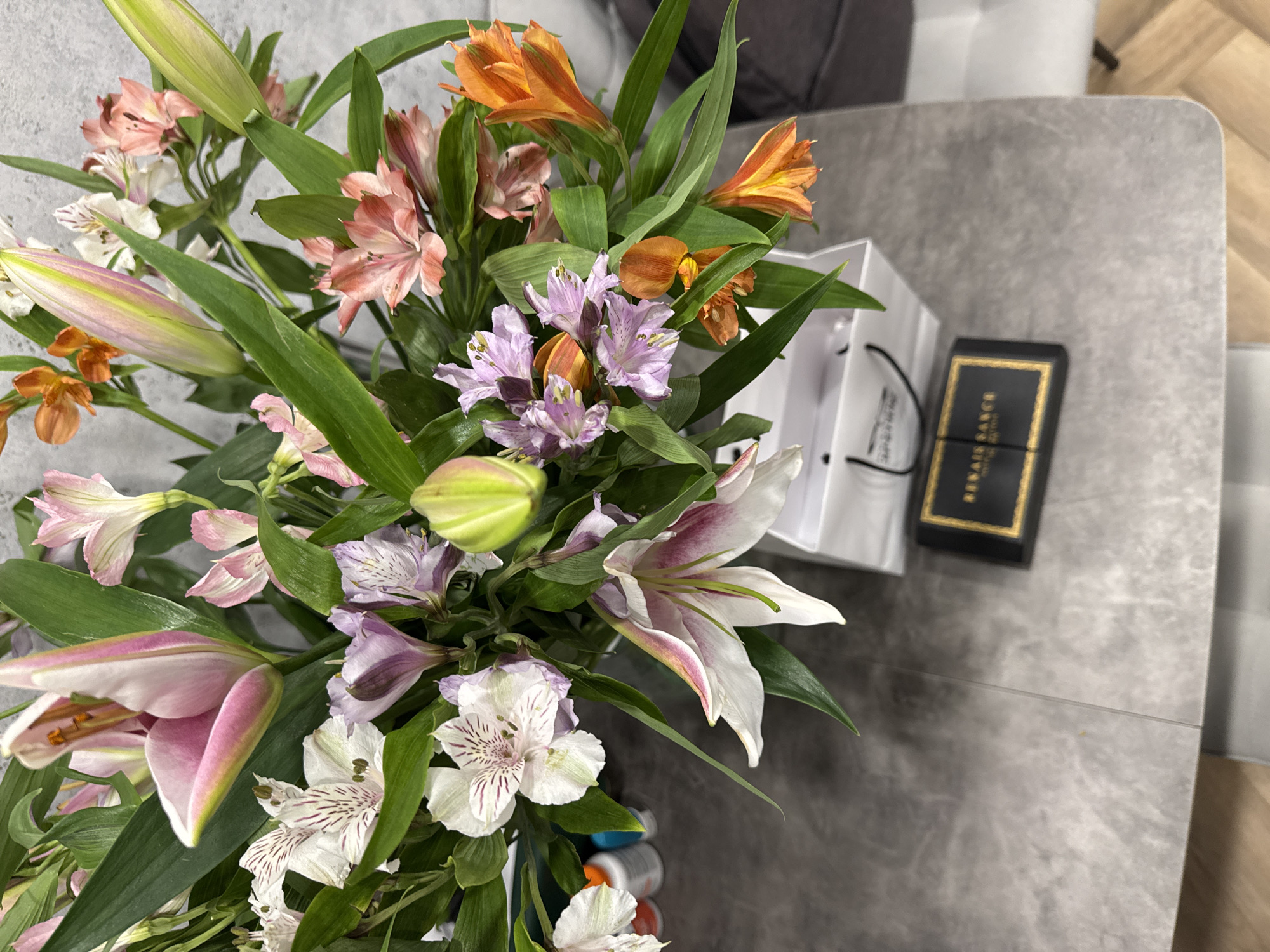 The image shows a bouquet of flowers on a grey table, with a white shopping bag and a black box next to it. The flowers are a mix of lilies and alstroemeria in various colors, including pink, white, orange, and purple. The black box has gold trim and text that reads "RENAN SANCE". The table appears to be in an indoor setting, possibly a home.