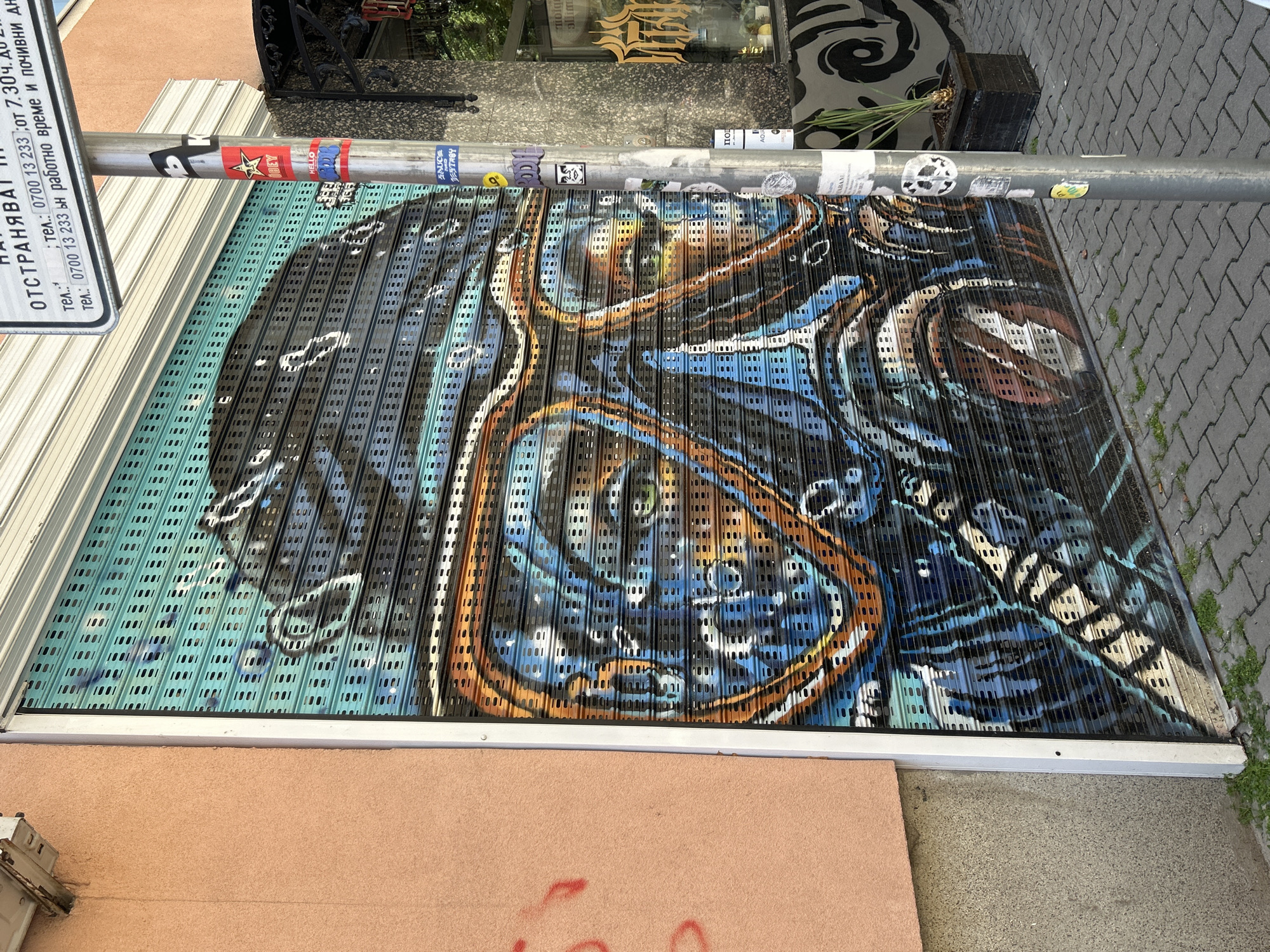 The image shows a metal roll-up security door with graffiti art. The artwork features a blend of blue, black, and orange hues. The door is located on a street in Unknown, Unknown, as can be inferred from the absence of definitive location markers. A metal pole covered in stickers stands adjacent to the door. A sign with Bulgarian text is affixed to the pole. The building's facade is painted in peach color. The ground is covered with gray paving stones. The artwork is abstract and occupies the majority of the door's surface.