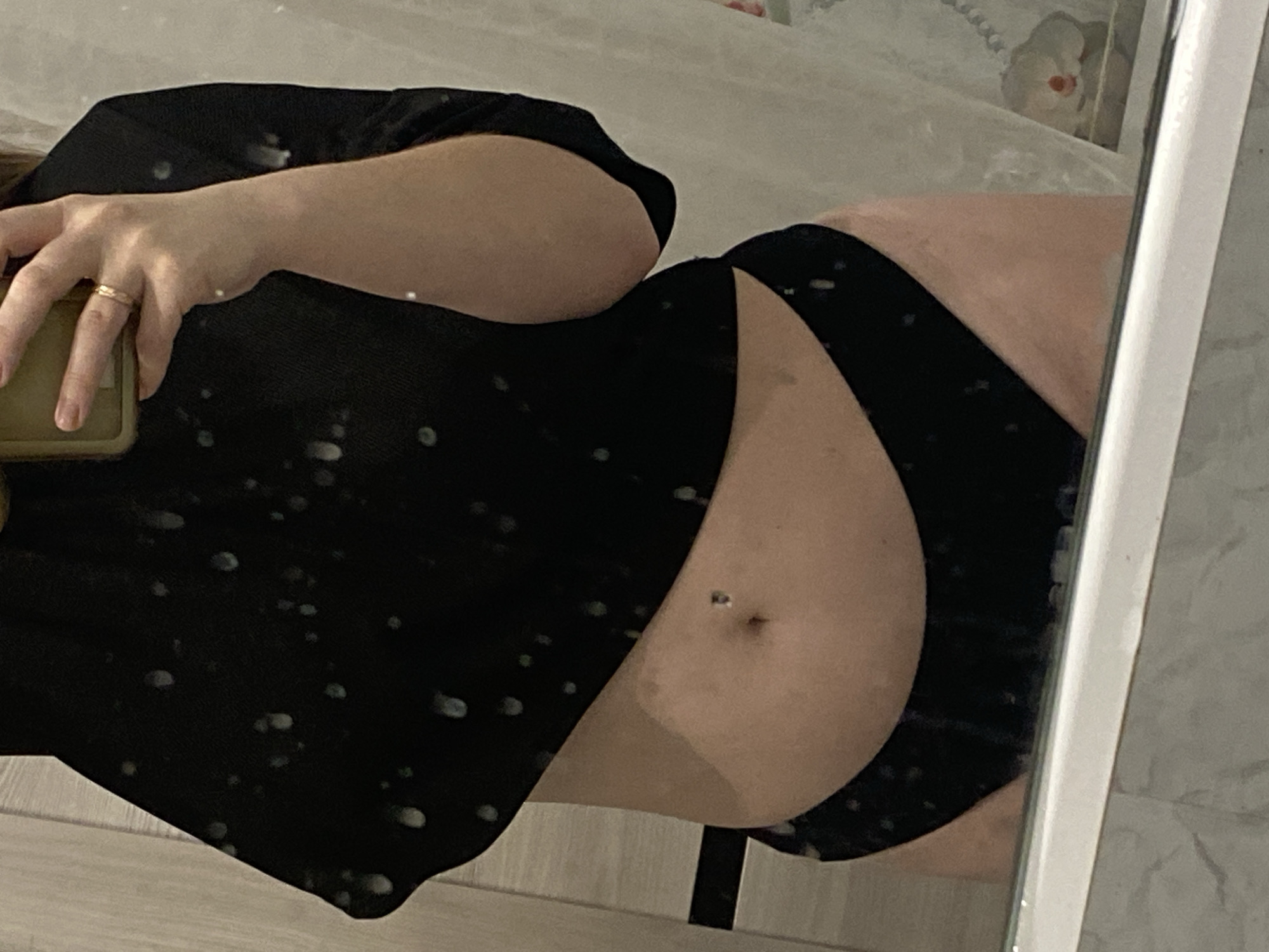 The scene depicts a mirror reflection of an individual's torso and partial arms, captured indoors within what appears to be a residential bathroom in the city of Noviy, Russia. The person is wearing a black cropped top, exposing the midriff, and black briefs. A metallic navel piercing is visible. The left hand, adorned with a gold-toned ring on the ring finger, holds a smartphone encased in a light-colored cover, indicating a selfie action. The mirror surface is speckled with numerous water droplets. The background reflected in the mirror displays a light-toned wooden door or wall paneling with vertical lines on the left, and a white or light-patterned shower curtain or opaque partition on the right. Below the primary reflection, a white horizontal fixture, consistent with a bathtub rim or vanity edge, is visible above a grey tiled surface.
