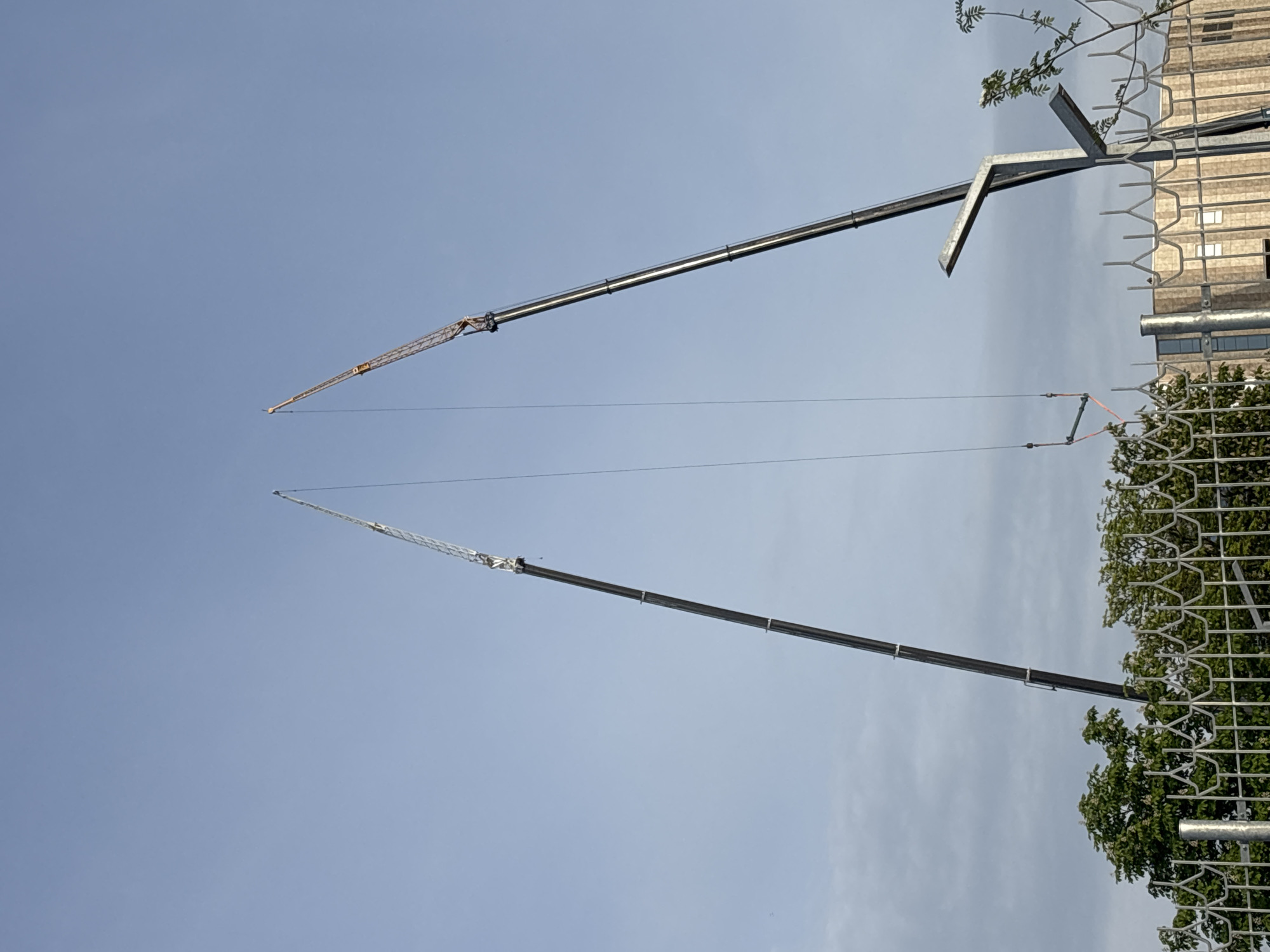 The image shows two tall cranes against a blue sky with some scattered clouds. The cranes are positioned diagonally, pointing towards the top of the frame. A metal fence and some trees are visible in the foreground on the right side. Behind the fence, there is a building with light-colored walls. The location is Minsk, Belarus.