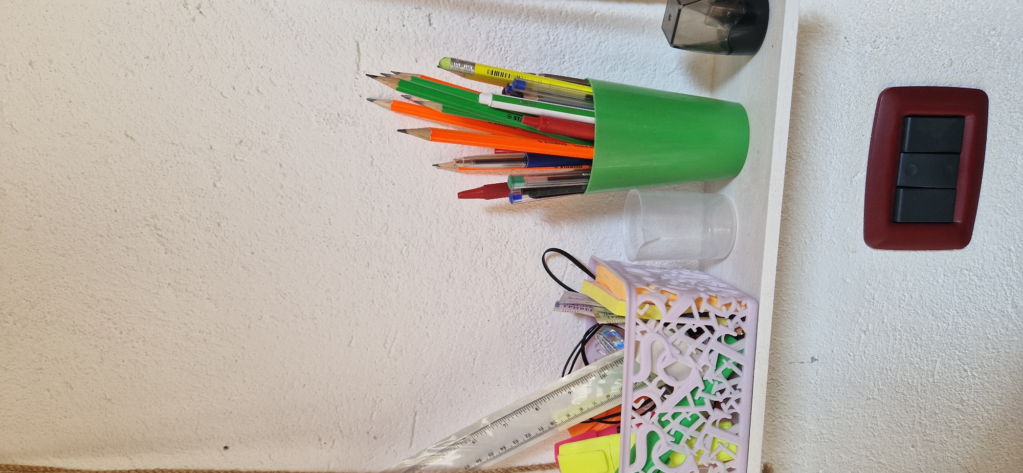 The image shows a close-up of a white, textured wall with a white shelf attached. On the shelf, there are several items, including a light purple basket filled with colorful stationery such as highlighters and pens. To the right of the basket, a green cup holds various pens and pencils. Further to the right, a small clear cup and a pencil sharpener are visible. Below the shelf, a dark red light switch with three black buttons is mounted on the wall. The overall scene appears to be an indoor setting, possibly a desk or study area. The image does not contain any specific landmarks or visual cues that would definitively place it in Brusson, Italy.