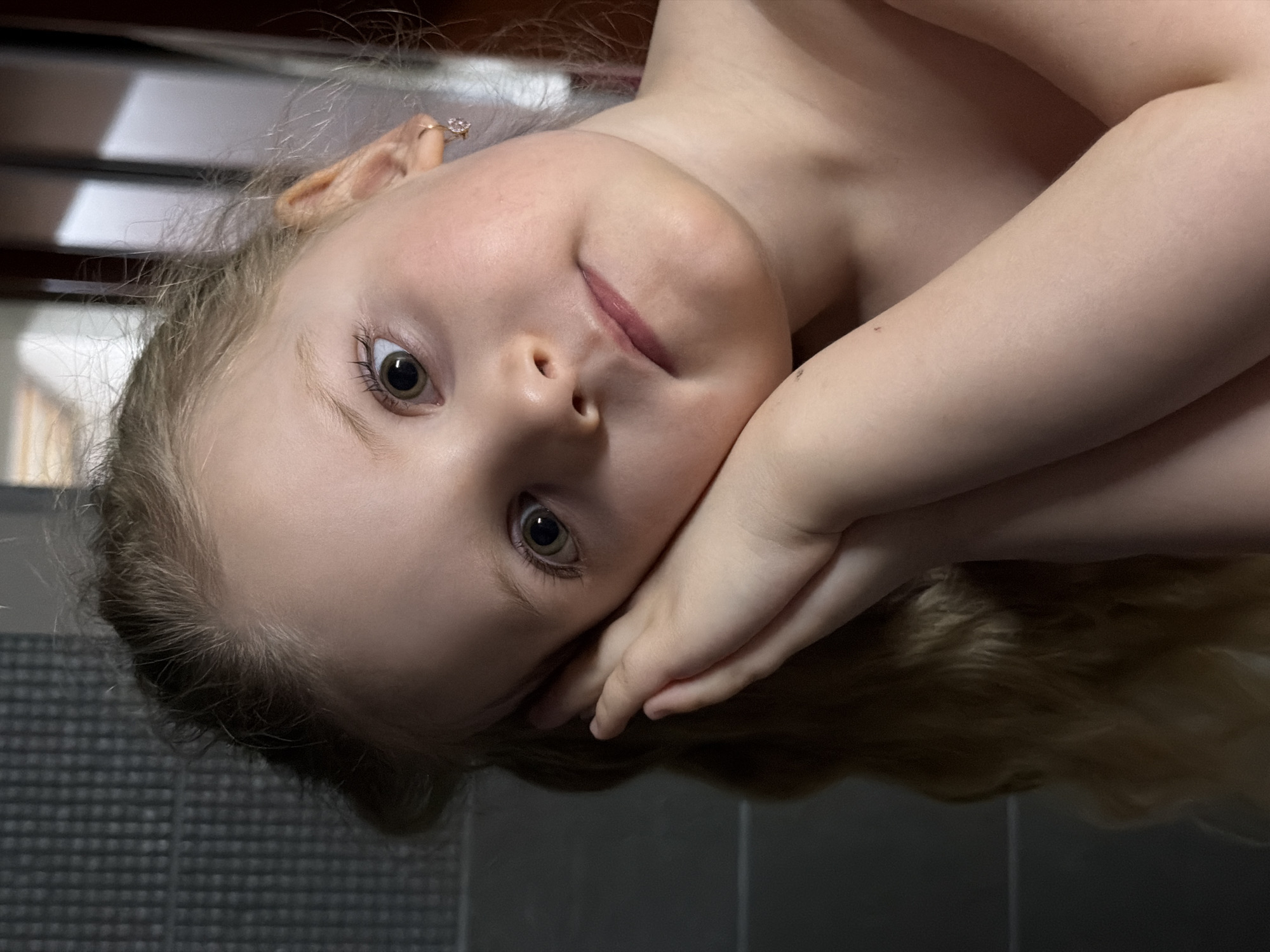 A young child with blonde hair and fair skin lies on her side, propped up by her arms. She is looking directly at the camera with a slight smile. The child has an earring on her left ear. The background appears to be a room with gray tiles on the floor. A staircase or another piece of furniture with white and dark wood is partially visible in the background. The location cannot be validated based on the image provided.