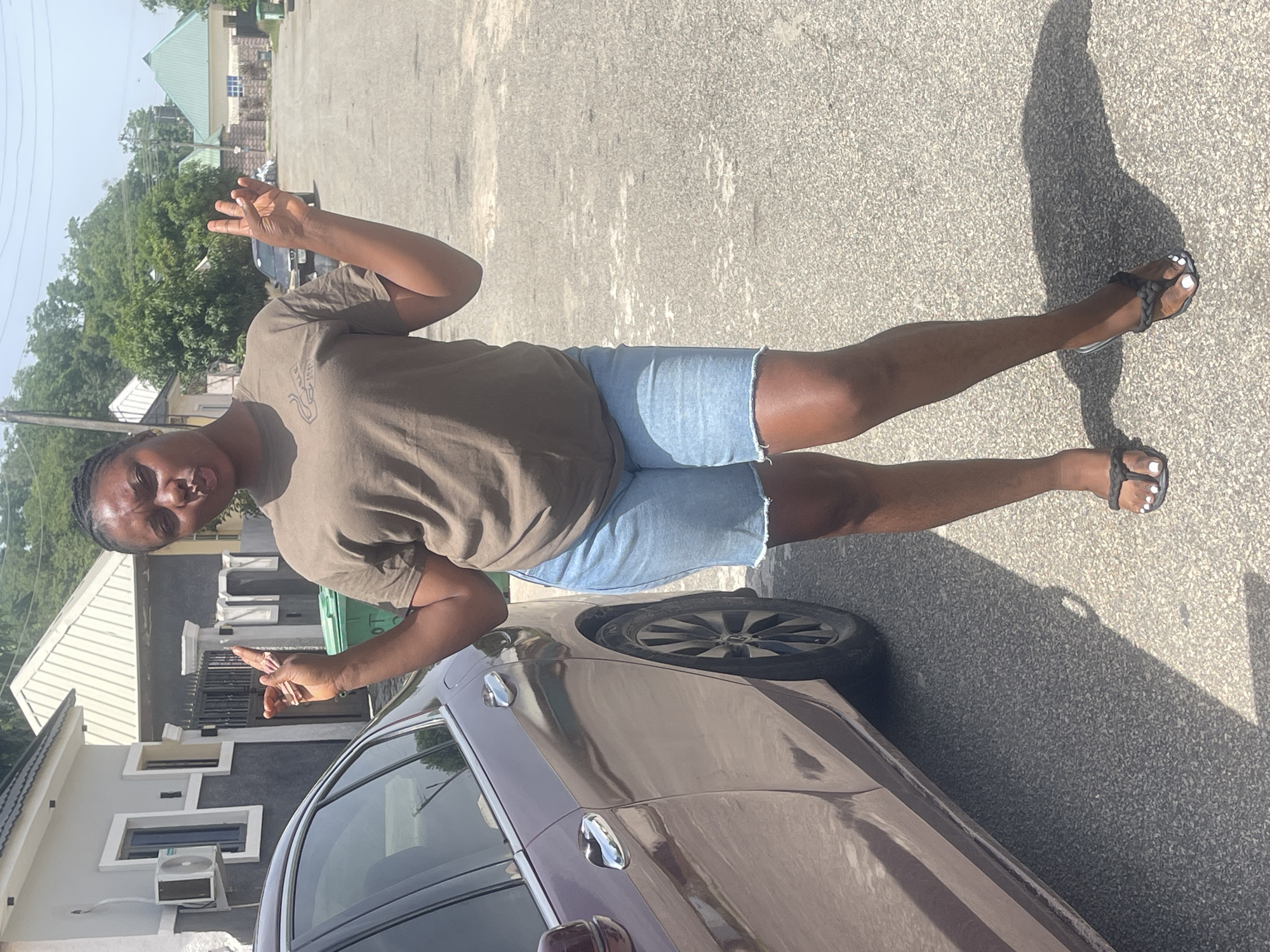 A woman is standing beside a car in an urban setting. She is wearing a brown shirt and blue jean shorts. She is making peace sign gestures with both hands. She is wearing black sandals with white nail polish. The car is brown, and a building with gray walls and a white roof is visible in the background. The setting appears to be in Abuja, Nigeria, based on architectural style. The ground is asphalt.