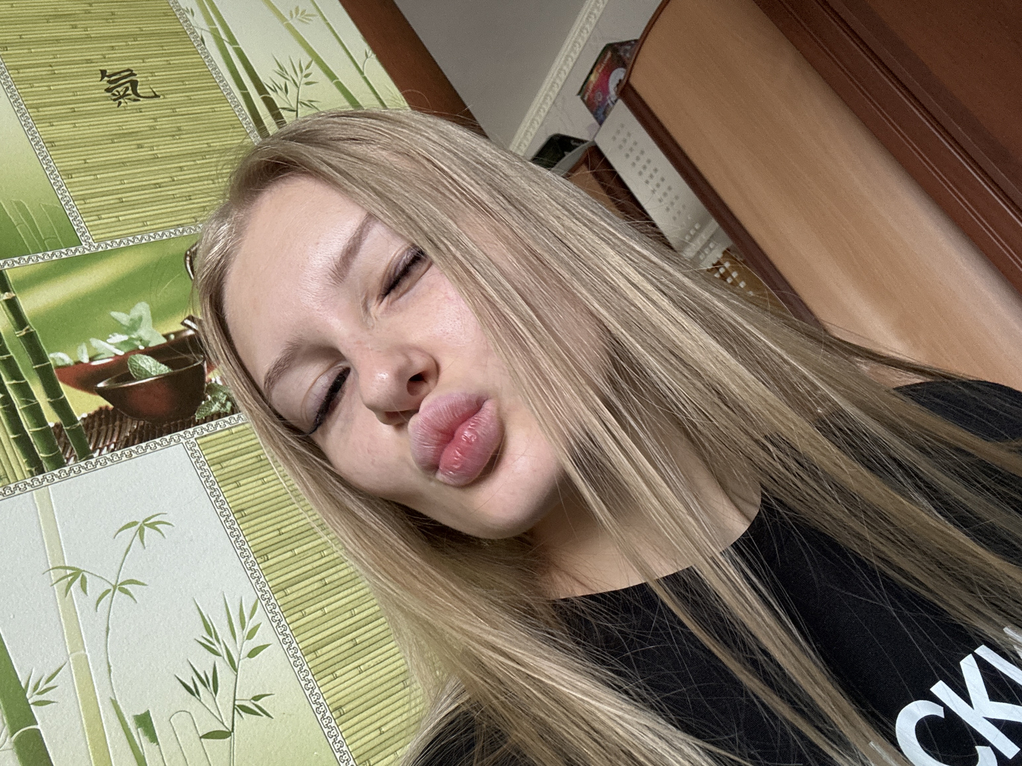 The image is a close-up, high-angle selfie of a young woman with long, blonde hair. She is wearing a black t-shirt and is puckering her lips towards the camera, with her right eye closed. The background appears to be a wall with a decorative pattern resembling bamboo. To the right of the woman, a wooden surface, possibly furniture, is visible. The lighting is moderate, casting soft shadows. The setting is indoors, likely a residential room. There is no discernible information in the image that would allow for the determination of its location within the city of Комарихинский, Russia.