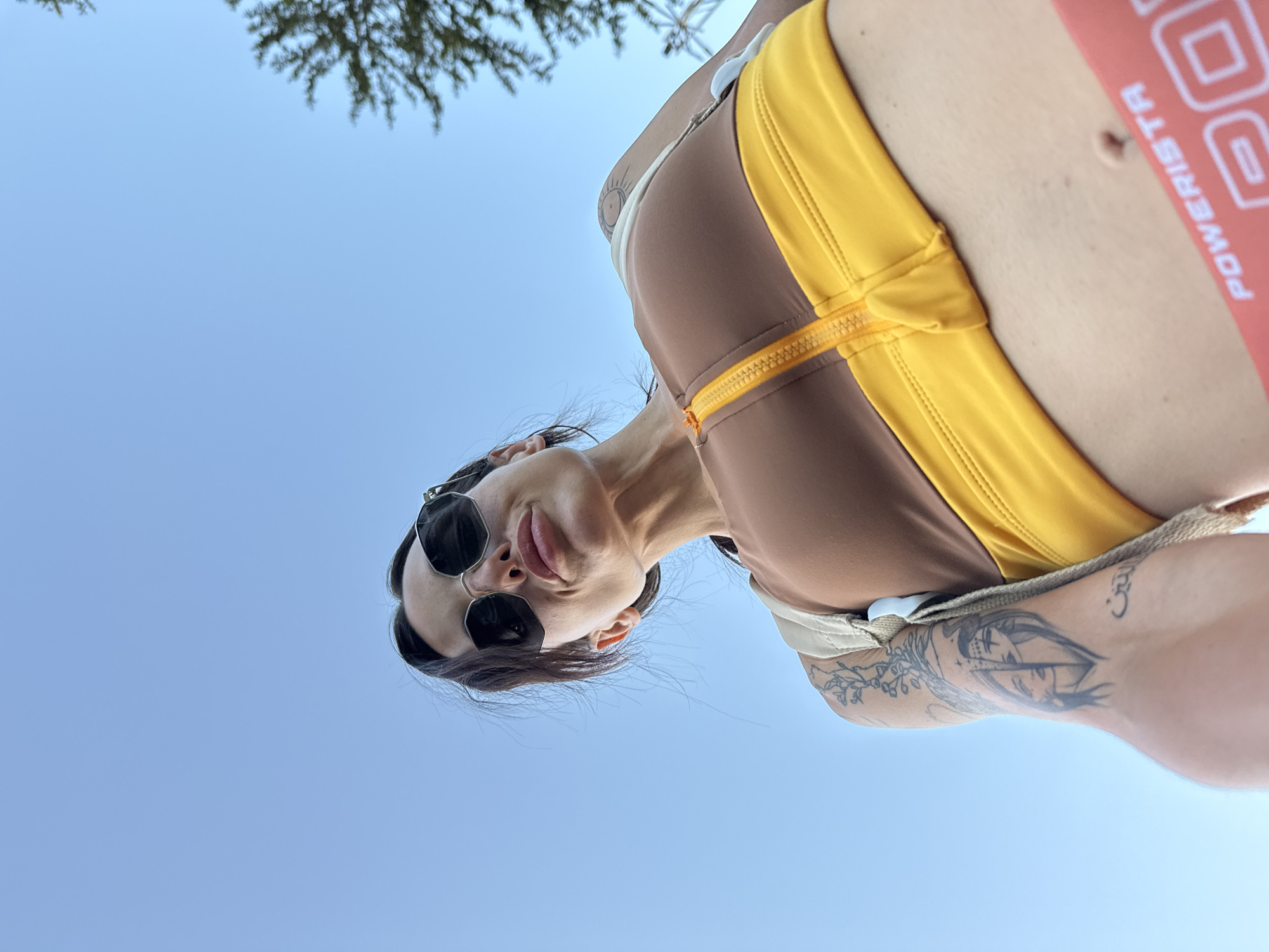 The image is a low-angle, close-up, outdoor selfie taken against a clear blue sky with the top of a tree visible on the right. The subject is a woman wearing oversized sunglasses, a brown and yellow zip-up athletic top, and red bottoms with the word "POWERISTA" printed on them. She has dark hair pulled back and visible tattoos on her left arm. The perspective suggests she is looking up at the camera. There are no discernible landmarks in the image to indicate a specific location within Pleven, Bulgaria.