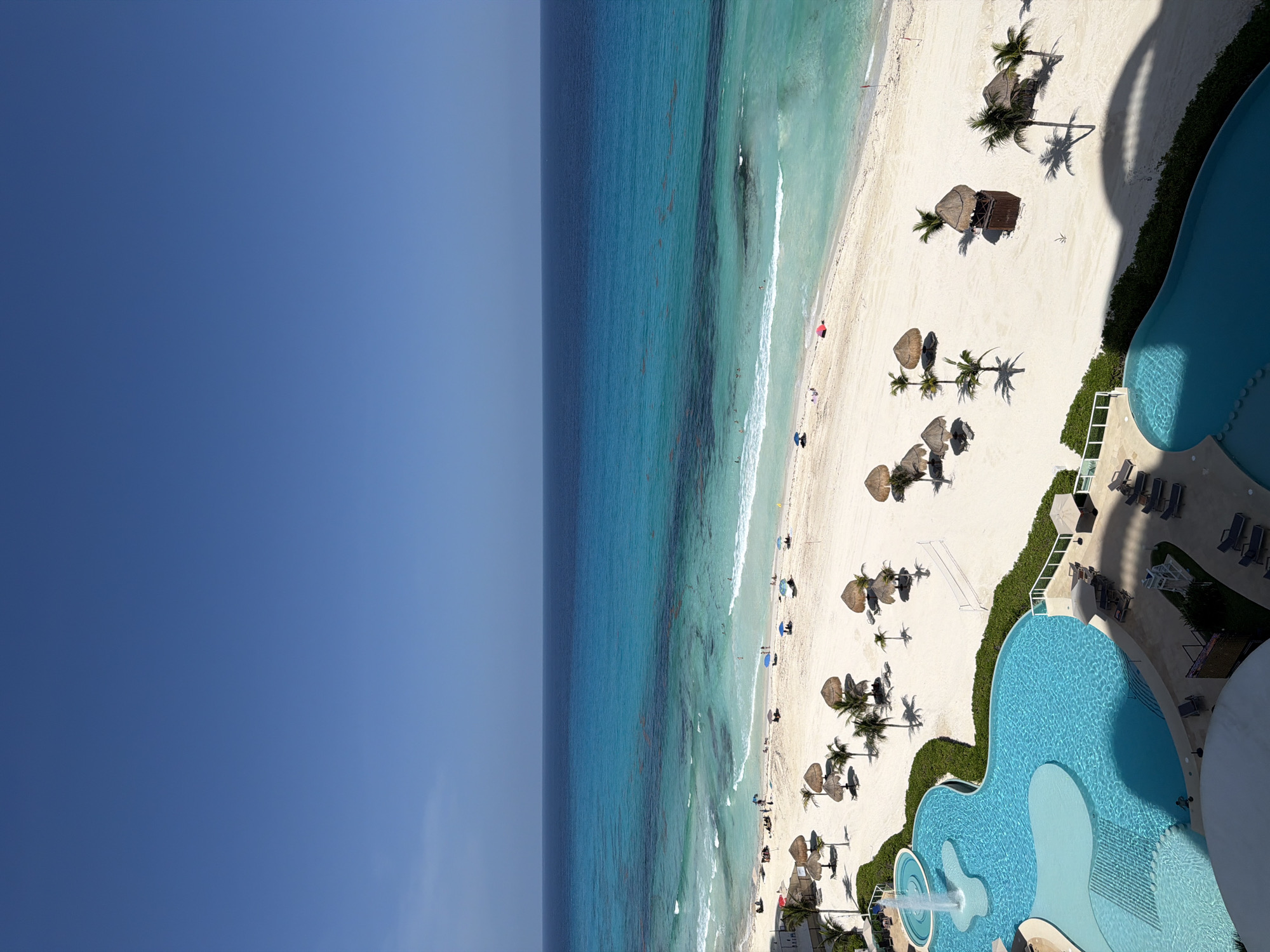 This high-angle, full-frame, vertical shot captures a sunny day at a beach resort in Cancún, Mexico. The scene is divided into three distinct horizontal bands: the clear, bright blue sky occupies the top third, transitioning to the deep blue of the ocean. The ocean itself showcases a gradient of turquoise and lighter blue hues, with visible seaweed or debris in the mid-distance. The foreground features a pristine white sand beach dotted with thatched palapa umbrellas and palm trees. Several lounge chairs are scattered across the sand, some occupied by people. A volleyball net is set up on the beach. To the left, a large, irregularly shaped swimming pool with a fountain is visible, adjacent to lush green landscaping. The edge of a building, presumably part of the resort, is seen at the bottom of the frame. The overall impression is one of a tropical vacation destination.