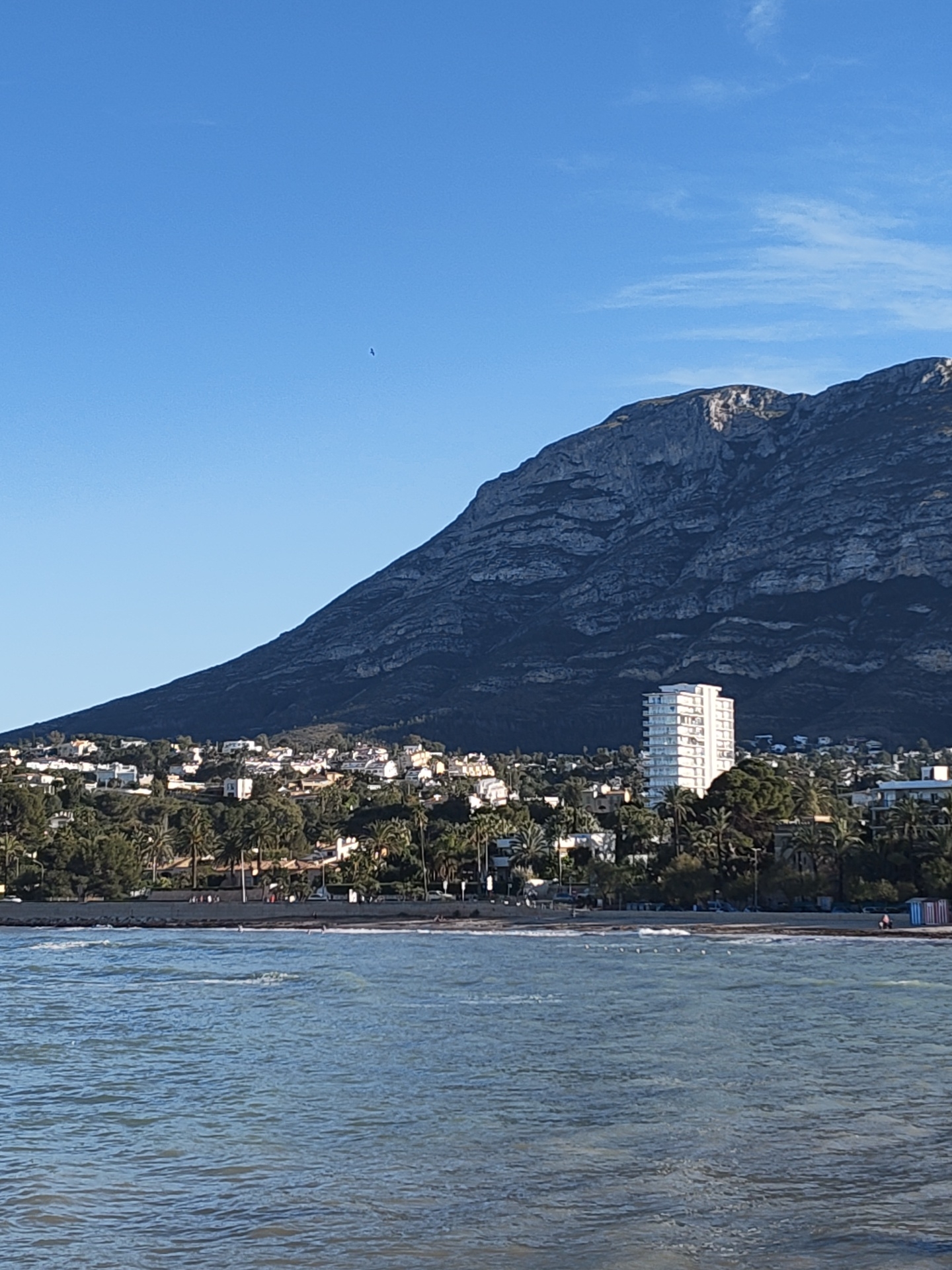The image shows a coastal view in Dénia, Spain. In the foreground is the Mediterranean Sea, with visible waves. Behind the sea, a beach is lined with sparse vegetation and a few indistinct figures, including a person on the far right and several unidentifiable objects. The middle ground features a cluster of buildings, including a prominent white multi-story structure, set among trees, likely a mix of palms and Mediterranean foliage. The backdrop is dominated by a large, rugged mountain with exposed rock formations, reaching high into a clear blue sky with a few scattered cirrus clouds.