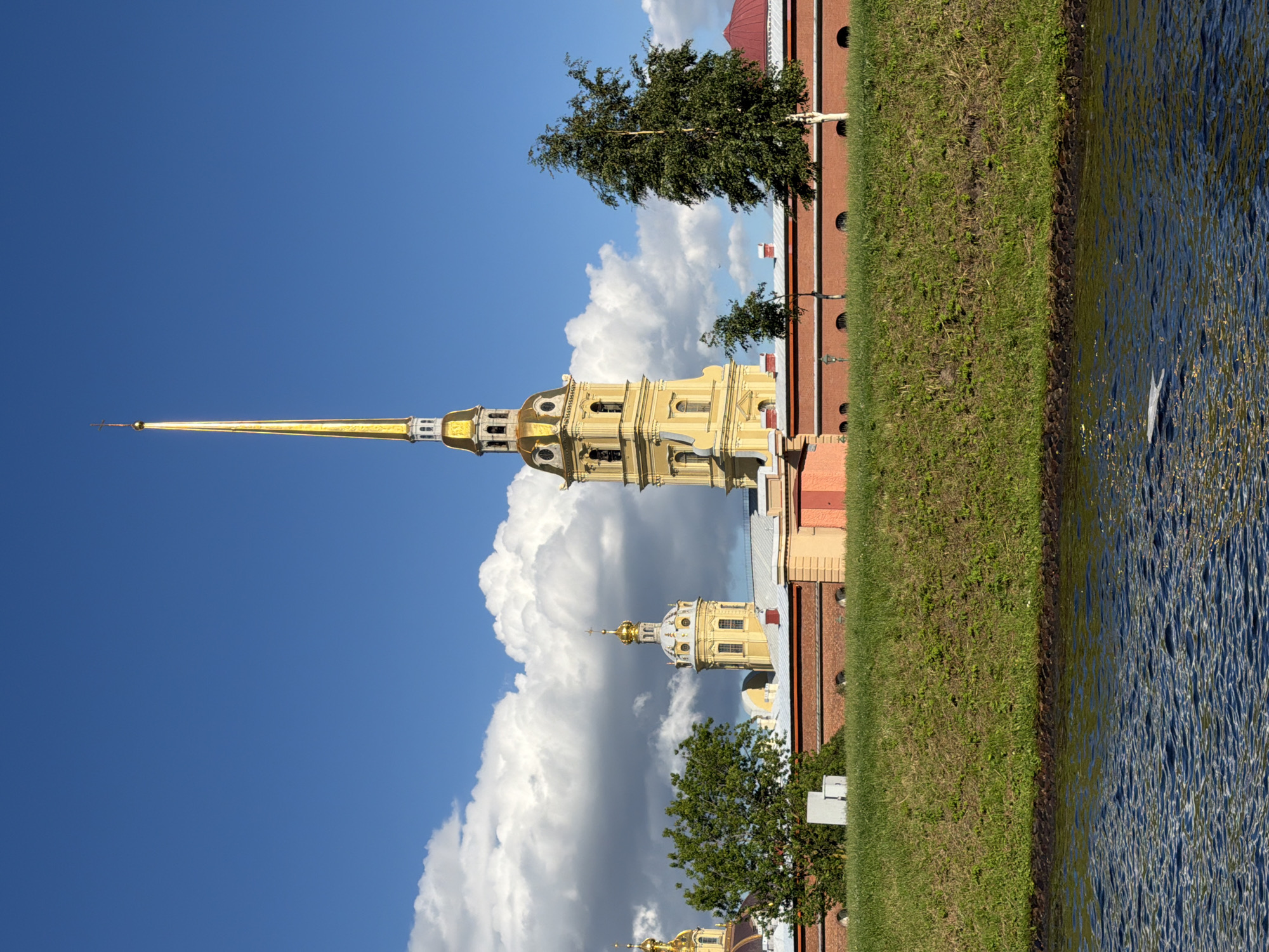 This is a vertical, eye-level, outdoor, full shot of the Peter and Paul Cathedral in St. Petersburg, Russia, taken on a sunny day with scattered clouds. The image is captured from a slightly elevated perspective, looking across a body of water towards the cathedral's iconic spire.
The main subject is the cathedral, a tall, yellow building with a golden spire topped by a cross. The spire is the most prominent feature, reaching towards the bright blue sky. Below the spire, the cathedral has multiple levels with arched windows and decorative elements. In the background, another smaller golden dome is visible. The building is bordered by a strip of green grass and then a body of dark blue water. The water's surface is rippled, suggesting a gentle breeze.
There are no people visible in the image. The weather appears to be pleasant, with a clear blue sky and fluffy white clouds. The sun is shining, casting bright light on the cathedral and the surrounding landscape. The time of day appears to be midday or early afternoon, given the brightness of the light. The location is identifiable as St. Petersburg due to the distinctive architecture of the Peter and Paul Cathedral. There is no visible text in the image.