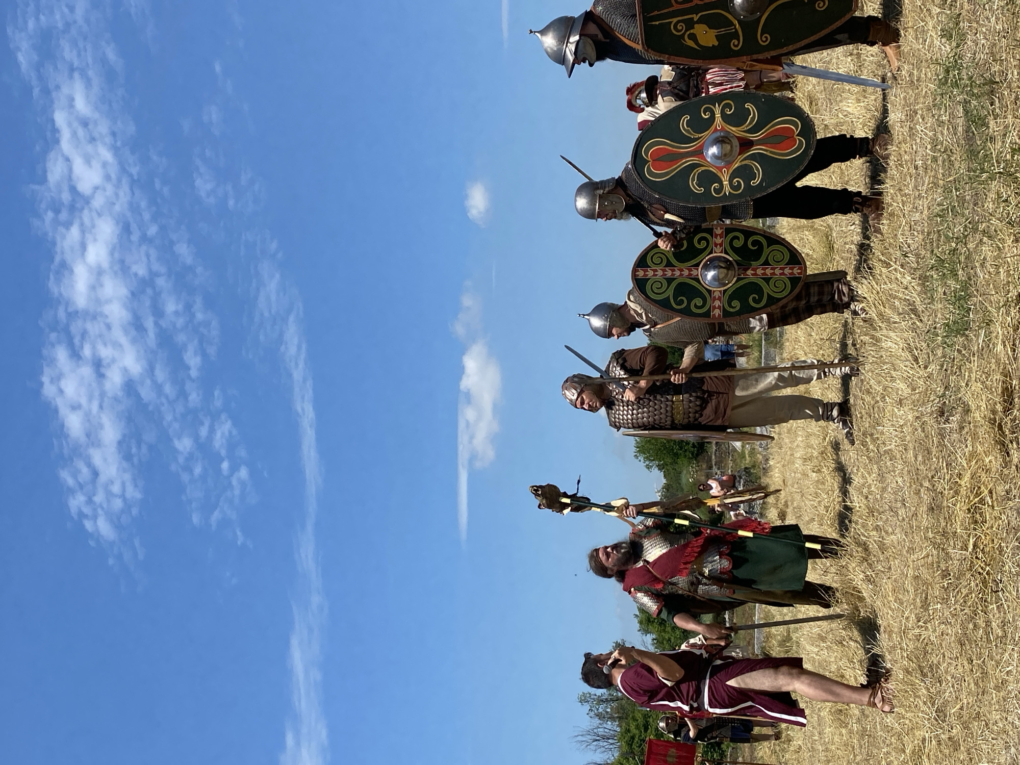 The image depicts an outdoor gathering of individuals in historical attire, set against a clear blue sky with scattered white clouds over Gigen, Bulgaria. The ground is covered in dry, golden-brown grass. Seven primary figures are arranged across the middle ground. On the left, a man wearing a maroon and white tunic, white skirt, and brown sandals, stands with bare legs, holding a dark, slender object near his mouth. Adjacent to him is a man with a full dark beard, clad in a green tunic, red sash, and scale armor, holding a tall standard topped with a bear's head, gazing upward and to the right. To the right of the standard-bearer, a man in scale armor over a brown tunic holds a vertical spear. Further to the right, three armored men stand side-by-side. Each wears a metal helmet, chainmail, and carries a sword, along with a large, round, dark green shield featuring red and green ornate patterns around a central boss. The shield on the far right also displays a yellow avian motif. In the background, sparse green trees, a red banner, and faint architectural elements are discernible.
