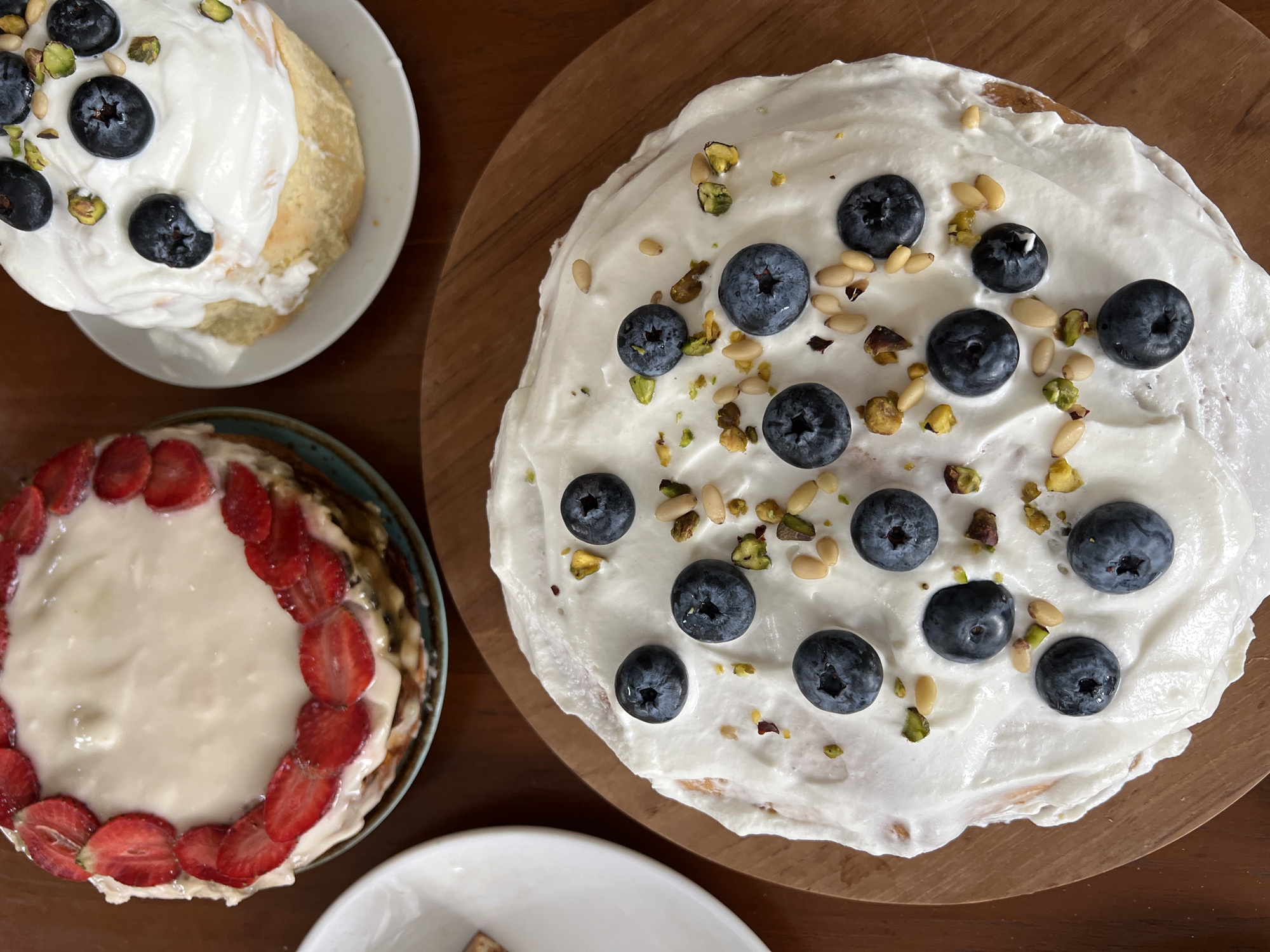 A high-angle, natural light shot shows three cakes on a wooden surface, possibly a table, in what appears to be a residential or dining area. The scene is likely located in Singakerta, Indonesia, based on provided context.
The largest cake is circular and sits on a wooden platter. It's topped with white frosting, blueberries, pine nuts, and chopped pistachios. To the upper left, a smaller cake or pastry is on a white plate, similarly topped with white frosting, blueberries, and nuts. Below left, another circular cake on a blue plate is garnished with sliced strawberries arranged around its perimeter, with a white filling in the center. The wooden surface has a warm tone and may be a table. Part of a white plate is also visible in the bottom center. The lighting is even, and the composition appears informal and candid.