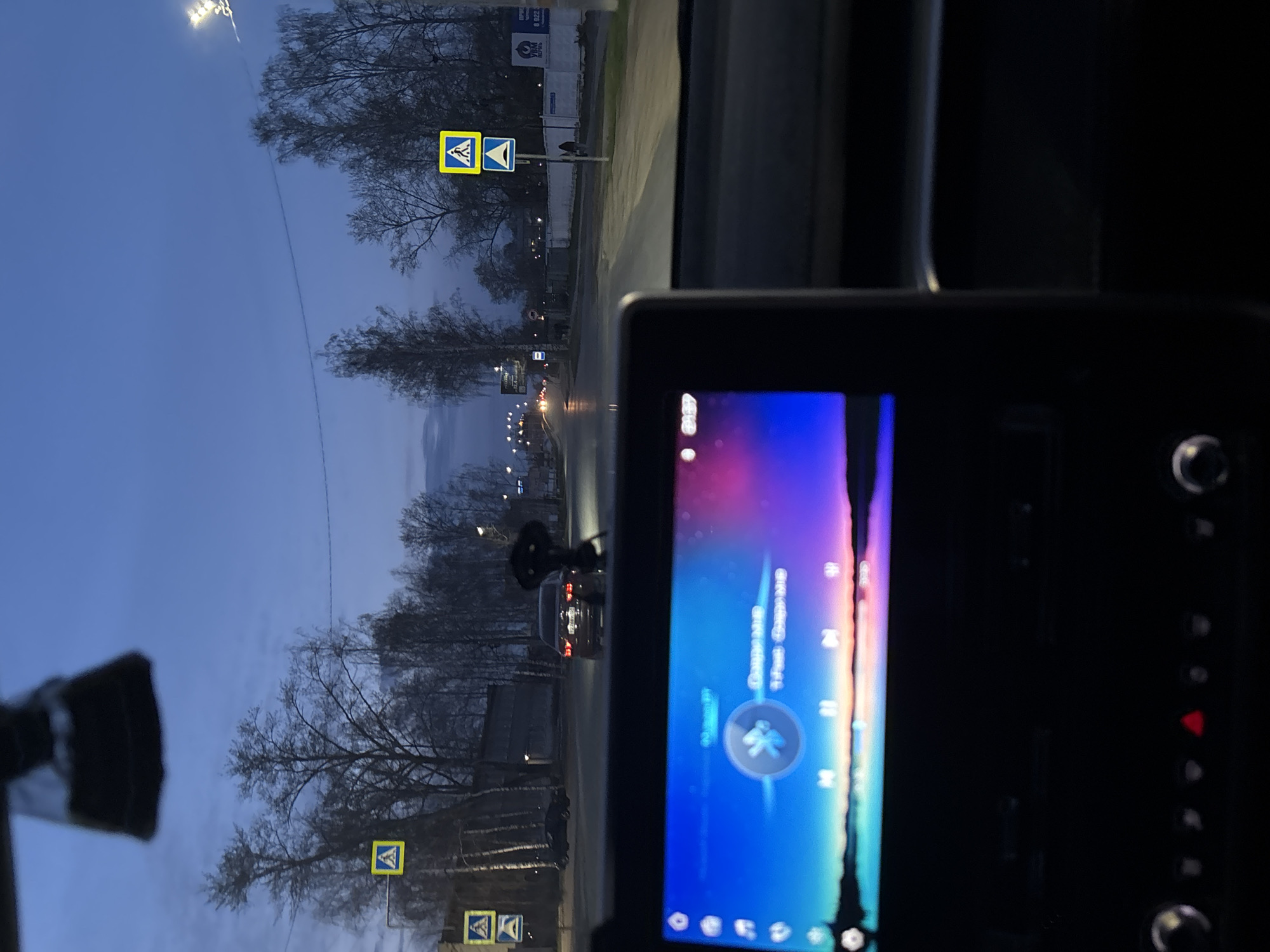 The photo was taken from inside a car on a street in Chaikovsky, Russia. The street appears wet, possibly after rain. Streetlights illuminate the scene. Several pedestrian crossing signs are visible along the road. A person is crossing the street ahead, close to a dark-colored car. In the foreground, a car's infotainment system is displaying music information.