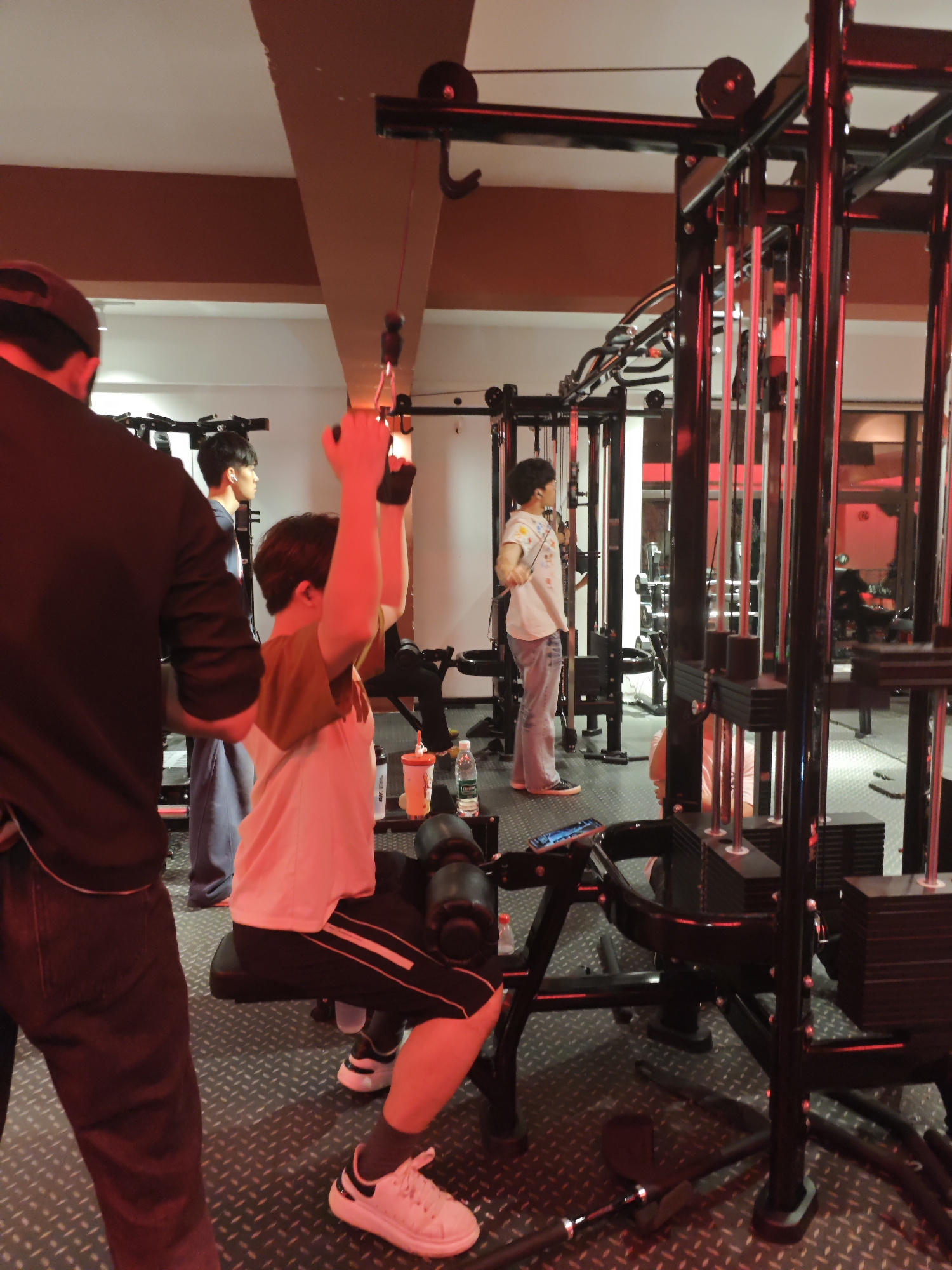 The image shows an indoor fitness center located in Chongqing High-tech Zone, China. A young individual is using a lat pulldown machine, with a trainer or spotter standing nearby. Another person is using a different machine in the background, and a third individual is standing further back, possibly observing or using equipment. The gym features standard weight machines, textured metal flooring, and red lighting. Drinks and a phone are placed on the machine near the person using the lat pulldown.