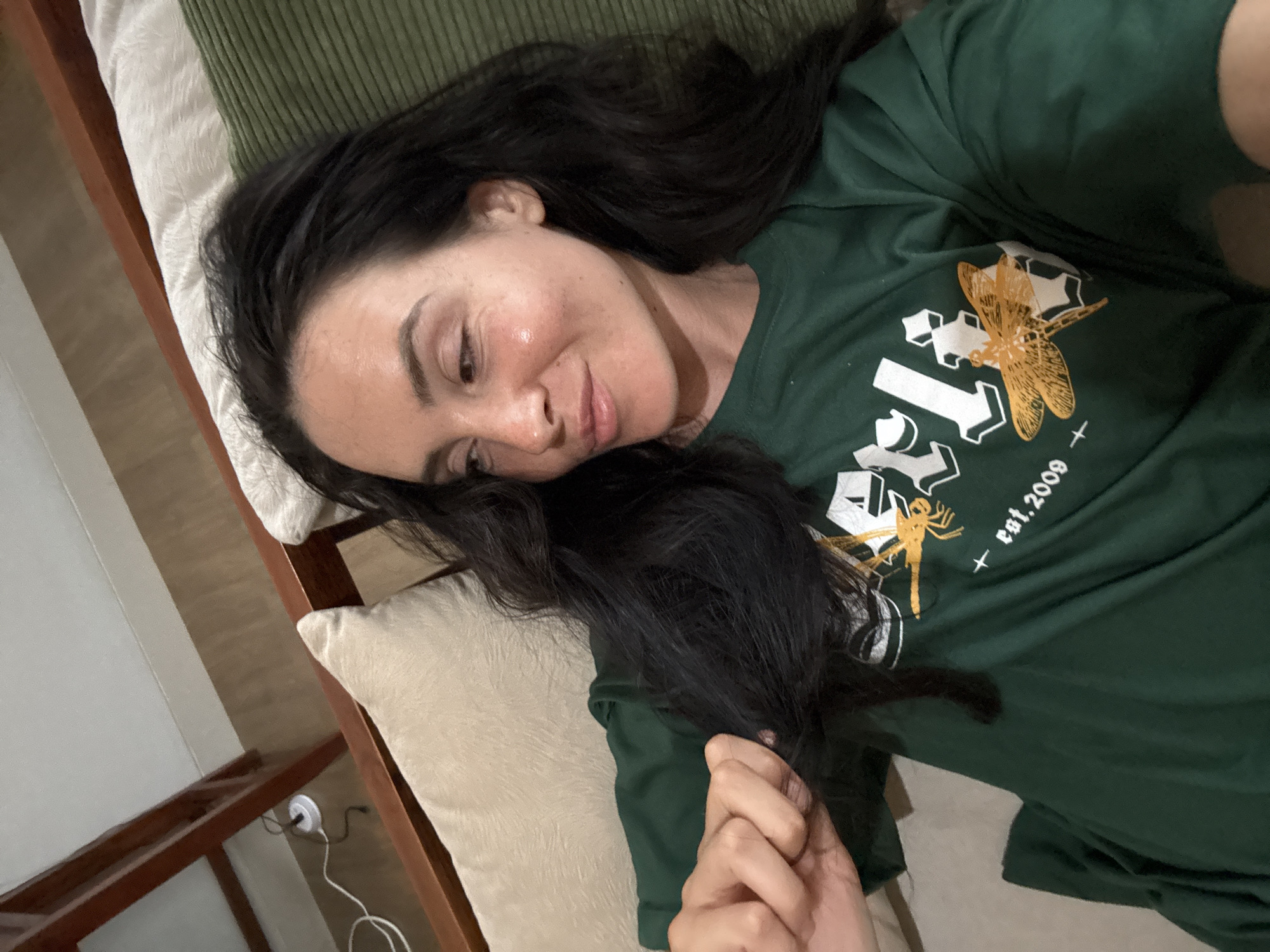 A woman with long, dark hair, wearing a green t-shirt, lies on a bed in a room. The room appears to be in North Kuta, Indonesia, based on the presence of wooden furniture and the style of the bed. The woman is looking to her right and touching her hair with her left hand. The bed has beige and green pillows. The background shows a wooden bed frame, a wall, and a floor. There is also an electrical cord plugged into a wall socket.