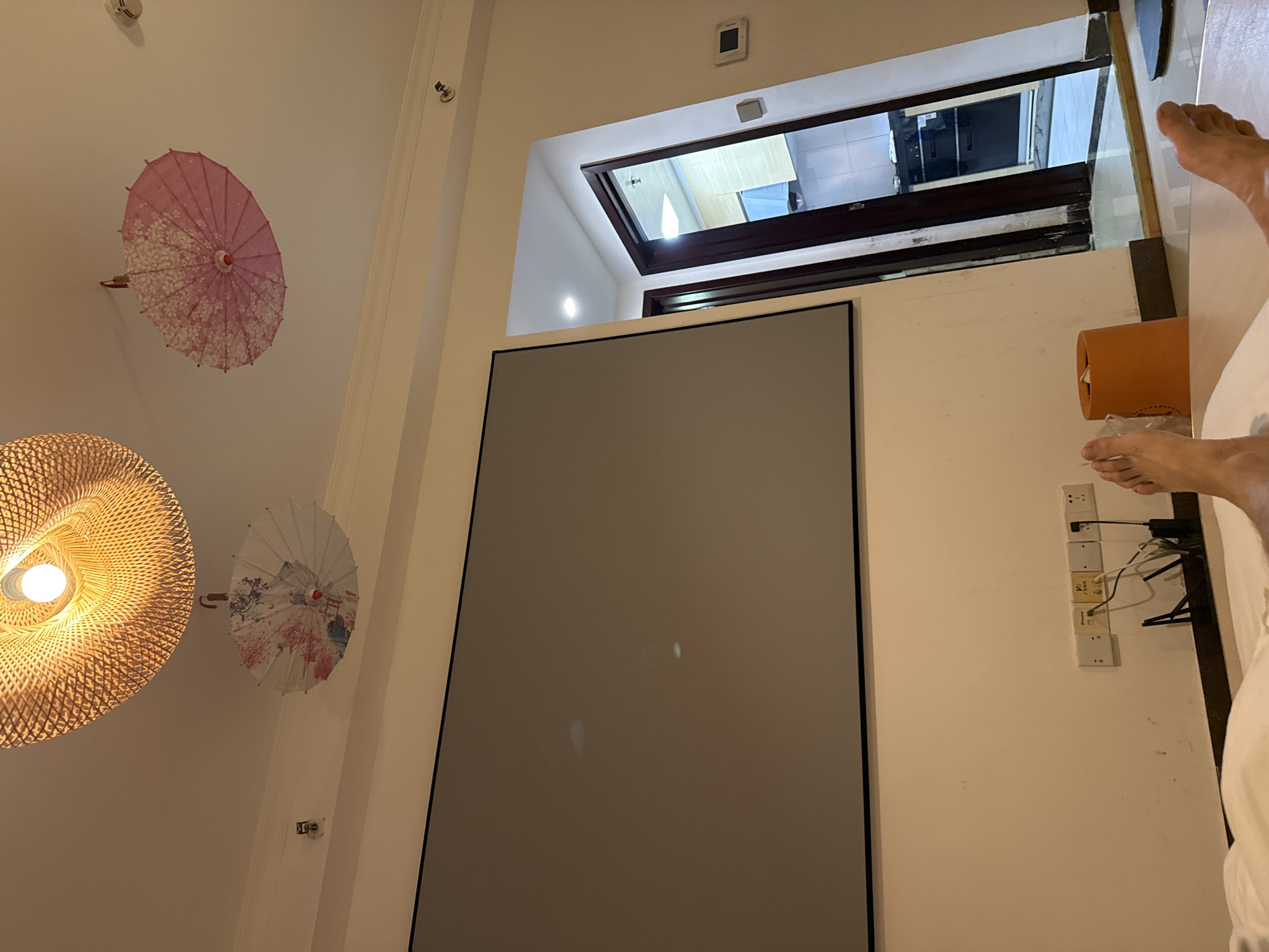The image depicts a room, possibly a bedroom or living area, with a large projector screen dominating the left side. The screen is gray and shows some faint, blurry light reflections. Above the screen, on the ceiling, are two decorative paper umbrellas, one pink with white floral patterns and another white with colorful floral designs. A woven light fixture with a visible light bulb hangs from the ceiling as well. To the right of the screen, a doorway leads into another room, which appears to be a kitchen or kitchenette, with light-colored cabinets and a dark countertop. In the foreground, the feet of a person are visible, lying down on what seems to be a bed or sofa, with a white sheet covering part of the lower body. Next to the feet is an orange cylindrical object, possibly a trash can or a decorative item. On the wall below the screen, there are electrical outlets and some cables plugged in. The overall impression is that of an indoor living space. The scene is not indicative of the Haizhu District of China, as there are no specific landmarks or elements that would place it there.