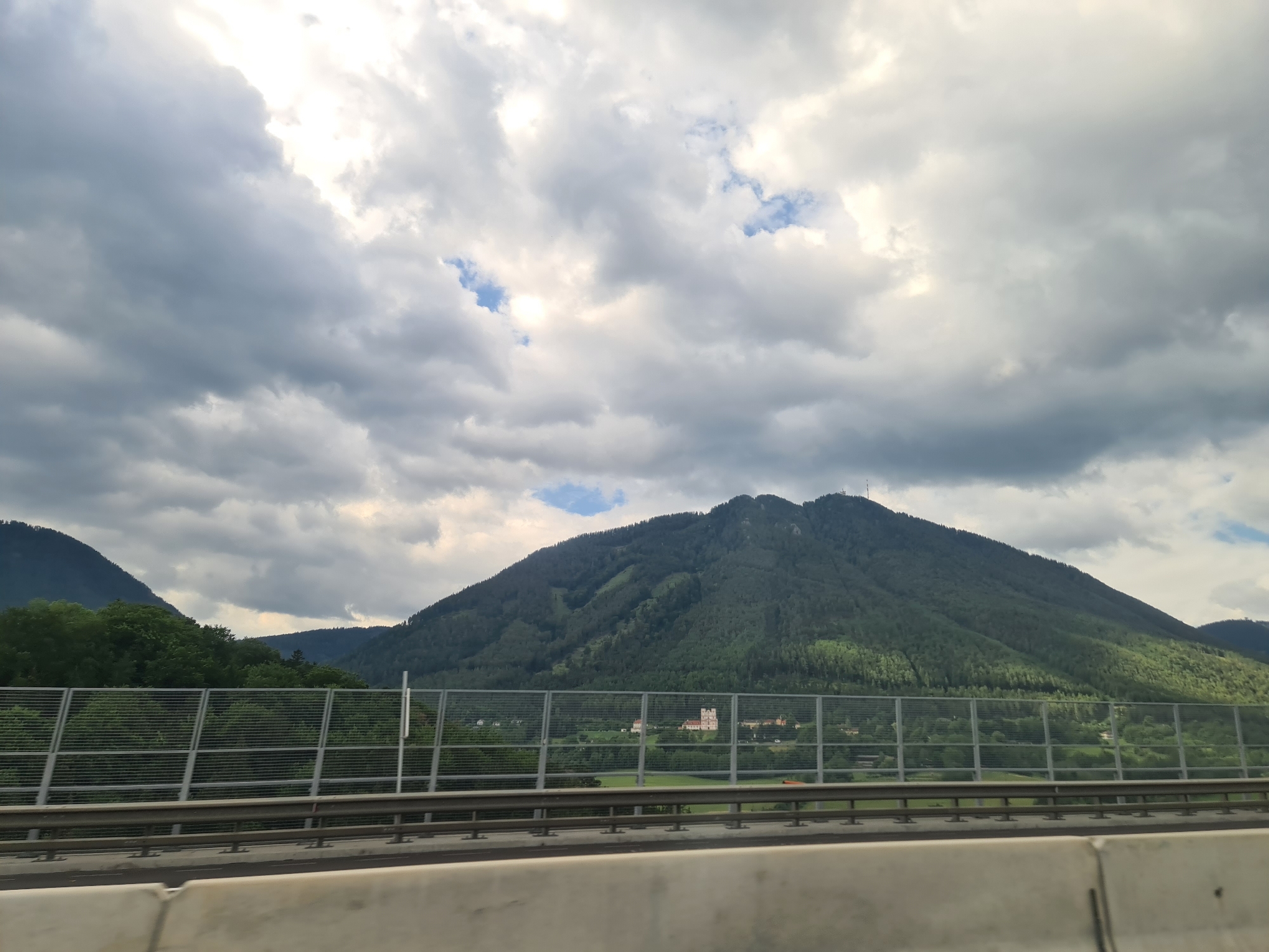 The image depicts a scenic view of a mountainous landscape with a cloudy sky, captured from a moving vehicle. In the foreground, a concrete barrier and a metal fence are visible, indicating the viewpoint is from a highway or elevated road. Beyond the fence, a green, forested mountain dominates the scene, with a building complex visible on its slope. The sky is filled with a mix of dark and light gray clouds, with patches of blue sky peeking through. The overall impression is one of a vast natural landscape under a dynamic sky. The location is identified as Schottwien, Austria, a town known for its mountainous terrain and picturesque views.