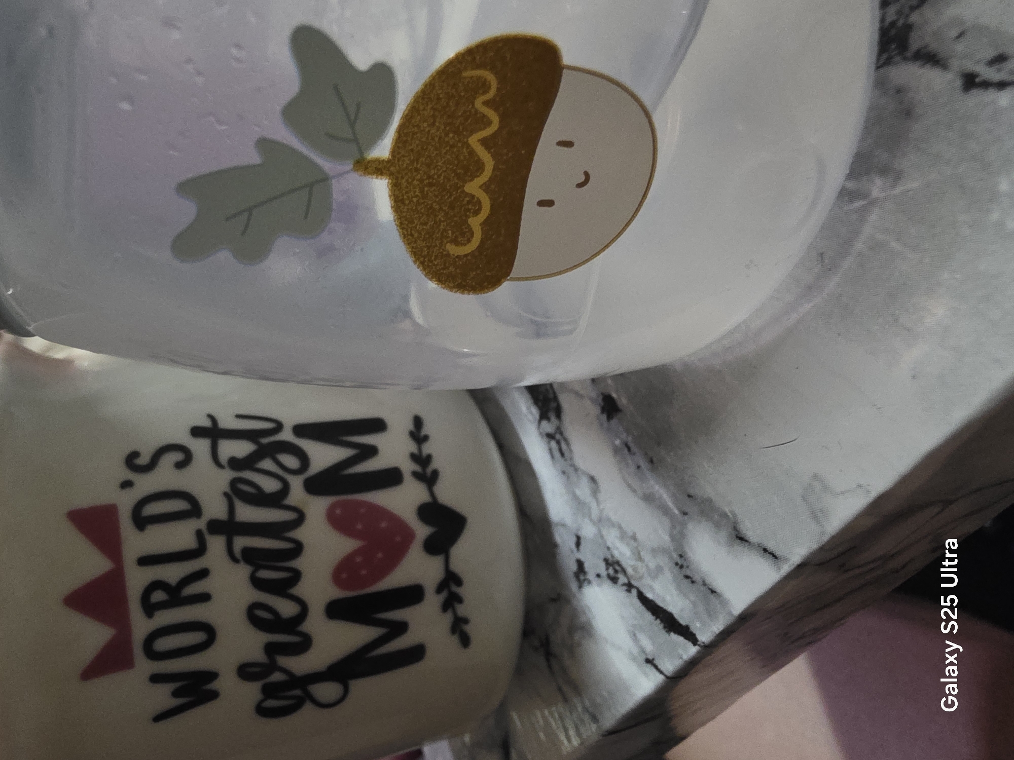 The image displays two cylindrical objects on a marble-patterned surface. The object on the left is a white mug with black text that reads "WORLD'S greatest MOM" and features a red crown and heart graphic. The object on the right is a clear plastic container with a cartoon acorn sticker on it, adorned with leaves. The text "Galaxy S25 Ultra" is visible in the bottom left corner of the image, suggesting it was taken with a Samsung Galaxy S25 Ultra phone. The setting appears to be indoors, on a countertop or table. There are no people visible in the image. The location in Cherencha, Bulgaria is not discernible from the visual content.
