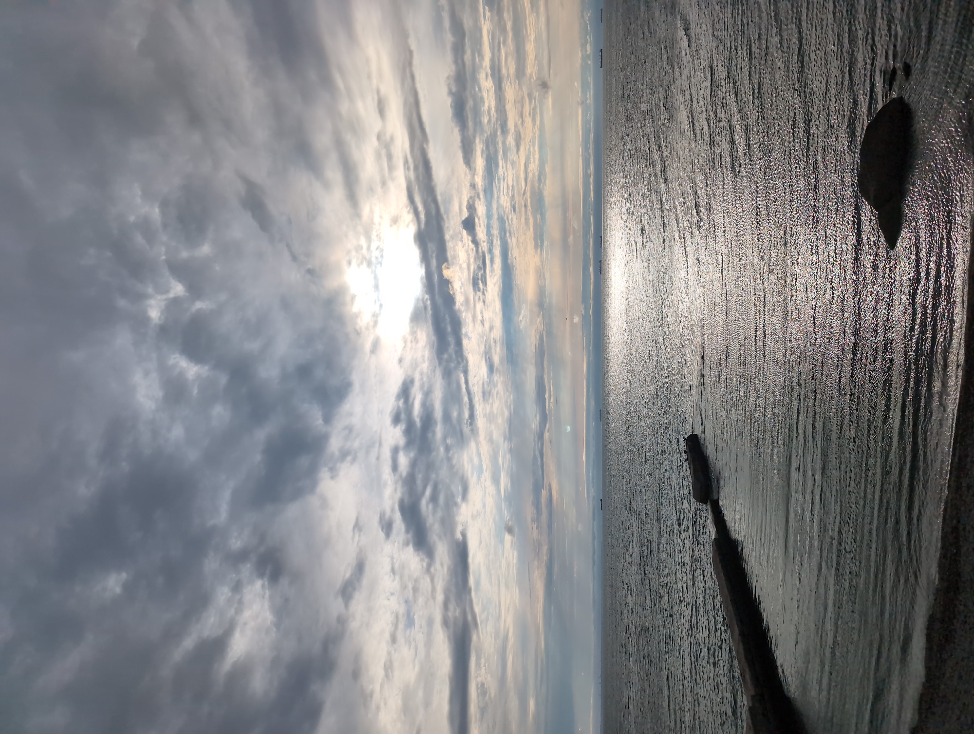 The image depicts a tranquil seascape with a dramatic cloudy sky. The sun is partially obscured by the clouds, casting a bright, shimmering reflection on the rippling surface of the water. In the foreground, a stone pier extends into the sea, with a large boulder visible near the shore. Several cargo ships are visible on the horizon, suggesting this is a view of the Black Sea near Odesa, Ukraine, possibly from the Odesa Port or a nearby coastal area. The overall atmosphere is one of calm and vastness, with the interplay of light and shadow creating a moody and atmospheric scene.